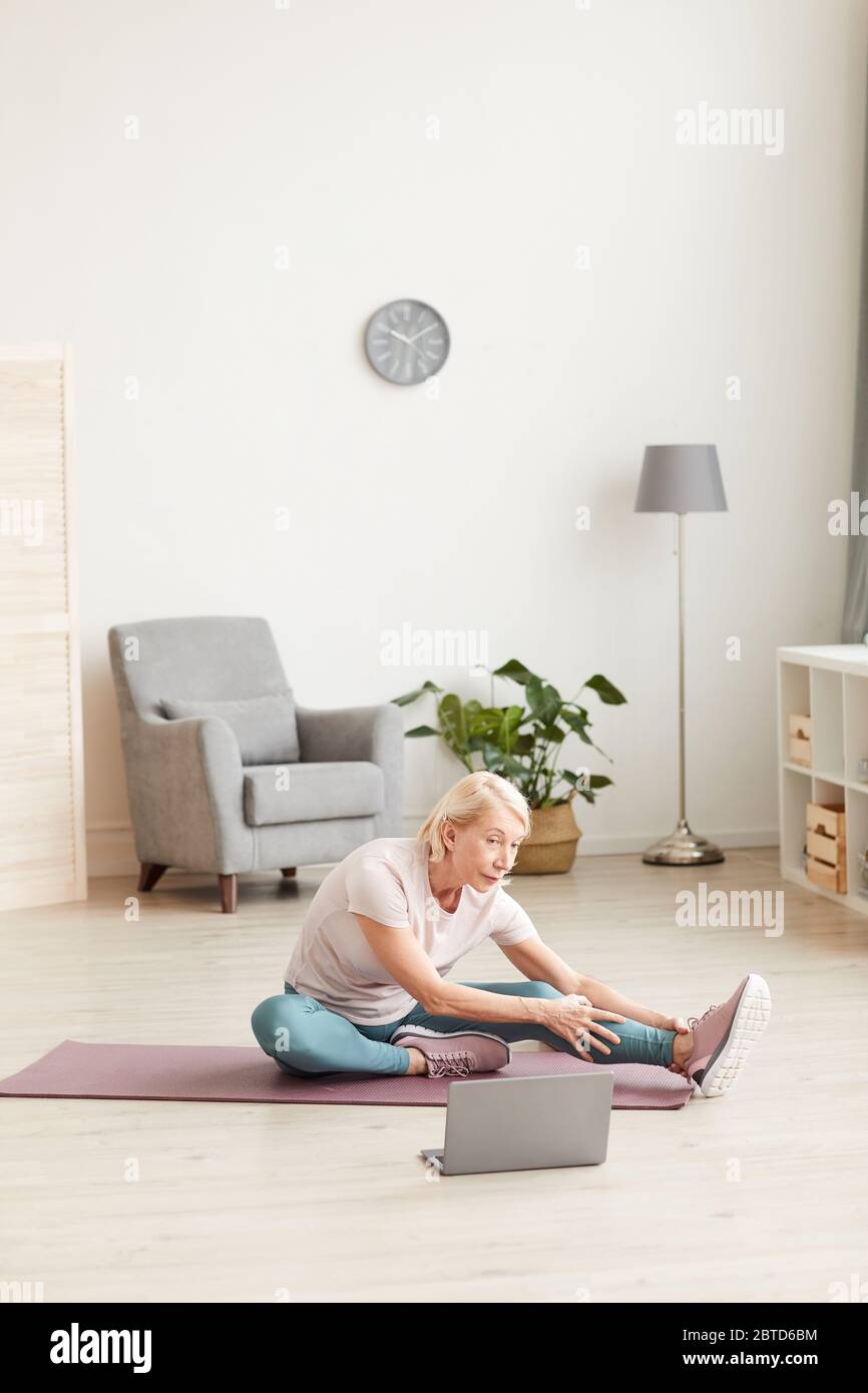 Ältere Frau sitzt auf Übungsmatte und streckt ihre Beine und schaut Online-Sporttraining auf Laptop zu Hause Stockfoto