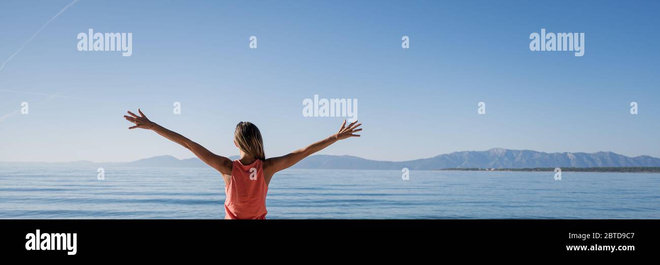 Blick von hinten auf eine junge Frau in rosa Kleidern, die am ruhigen Morgenmeer steht und ihre Arme weit gestreut hat, während sie den neuen Tag begrüßt. Weitwinkel ima Stockfoto