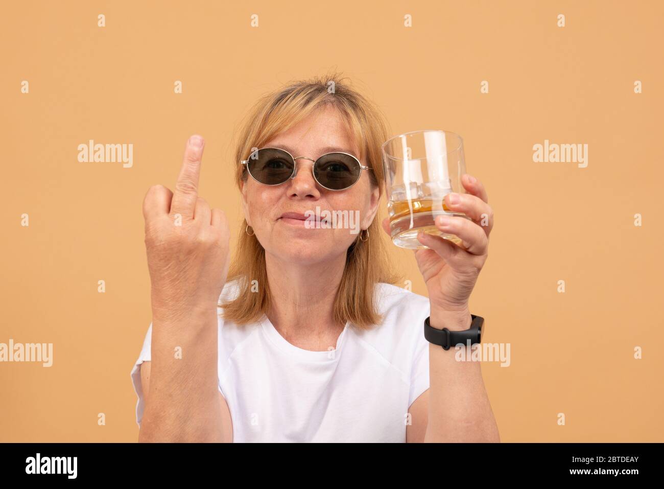 Frau mittleren Alters in schwarzer Sonnenbrille hält Glas Whisky in der Hand, zeigt Mittelfinger an der Kamera Stockfoto