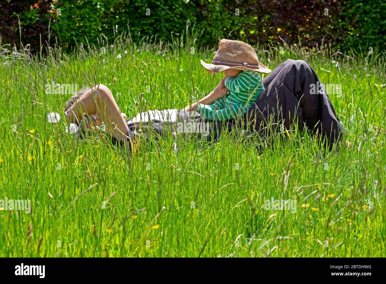 Person Großvater liegt in Wiese lange Gras Rasen hält Baby Tragen Strohhut im Frühling Mai Garten 2020 Carmarthenshire Wales UK KATHY DEWITT Stockfoto