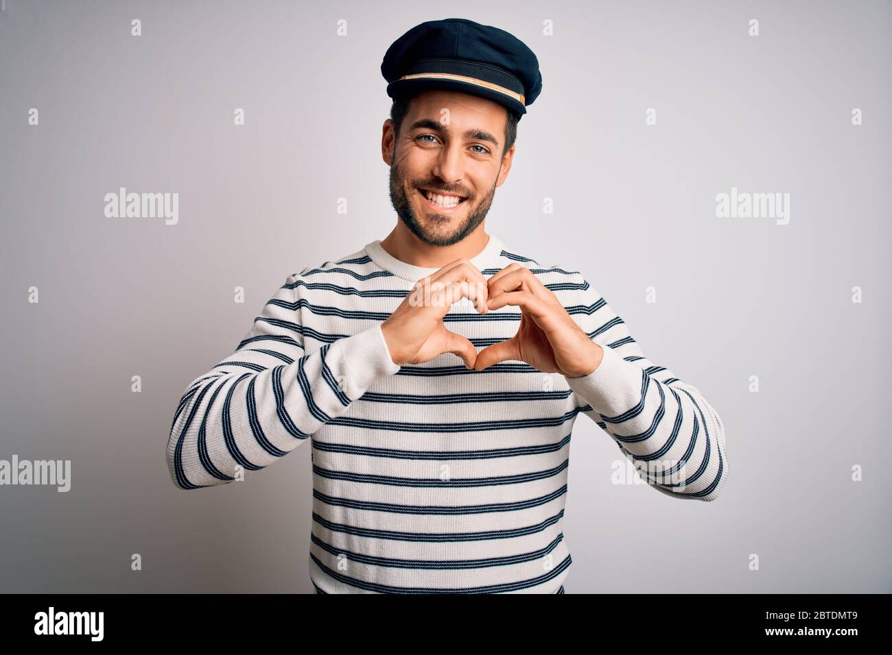 Junge schöne Seemann mit Bart trägt marineblau gestreifte Uniform und Kapitän Hut lächelnd in der Liebe zeigt Herz Symbol und Form mit den Händen. Romantisch Stockfoto