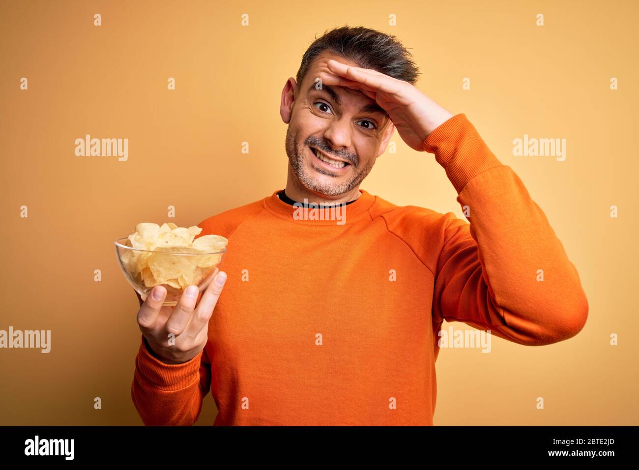 Junger schöner Mann hält Schüssel mit Kartoffeln Chips über isoliert gelben Hintergrund gestresst mit Hand auf Kopf, schockiert von Scham und Überraschung Gesicht, Stockfoto