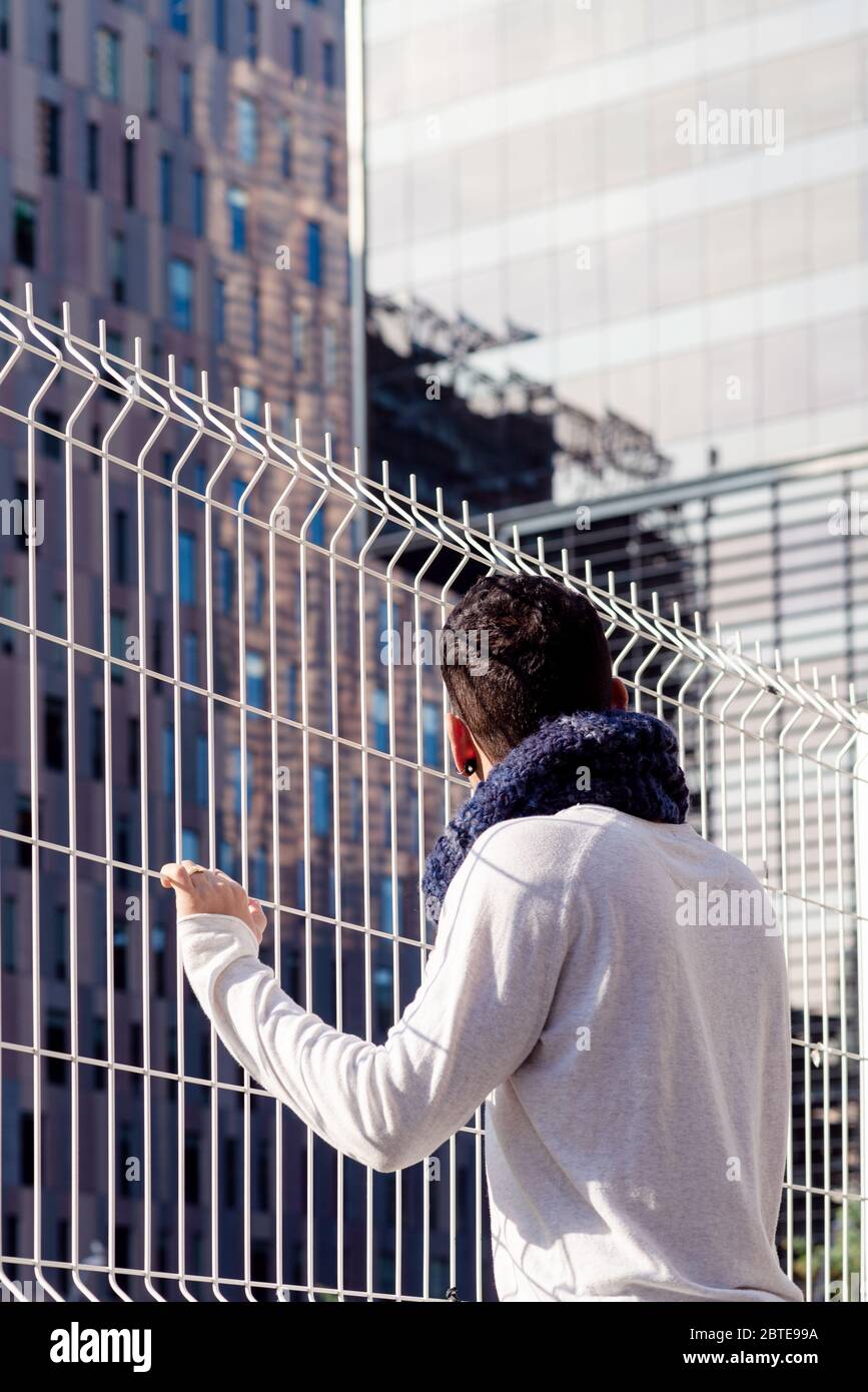 Junger Mann, der sich auf einen Metallzaun lehnt und auf die Geschäftsgebäude blickt Stockfoto