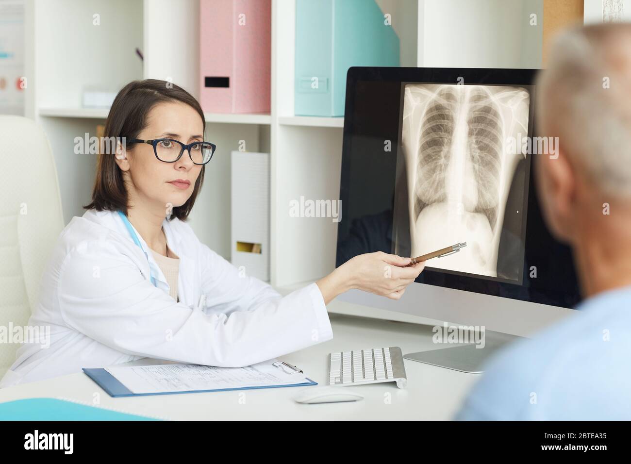 Porträt einer ernsthaften Ärztin, die auf das Röntgenbild von Lunge und Brust zeigt, während sie die leitende Patientin in der Klinik konsultiert Stockfoto