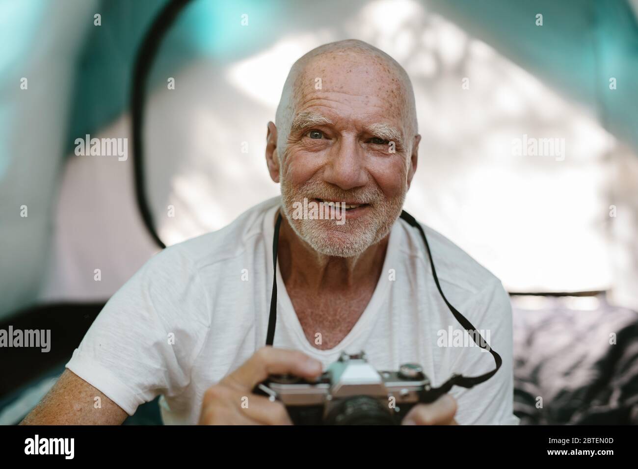 Nahaufnahme eines älteren Mannes, der mit einer Digitalkamera vor seinem Zelt sitzt. Pensionierter Mann fotografiert während des Campens in der Natur. Stockfoto