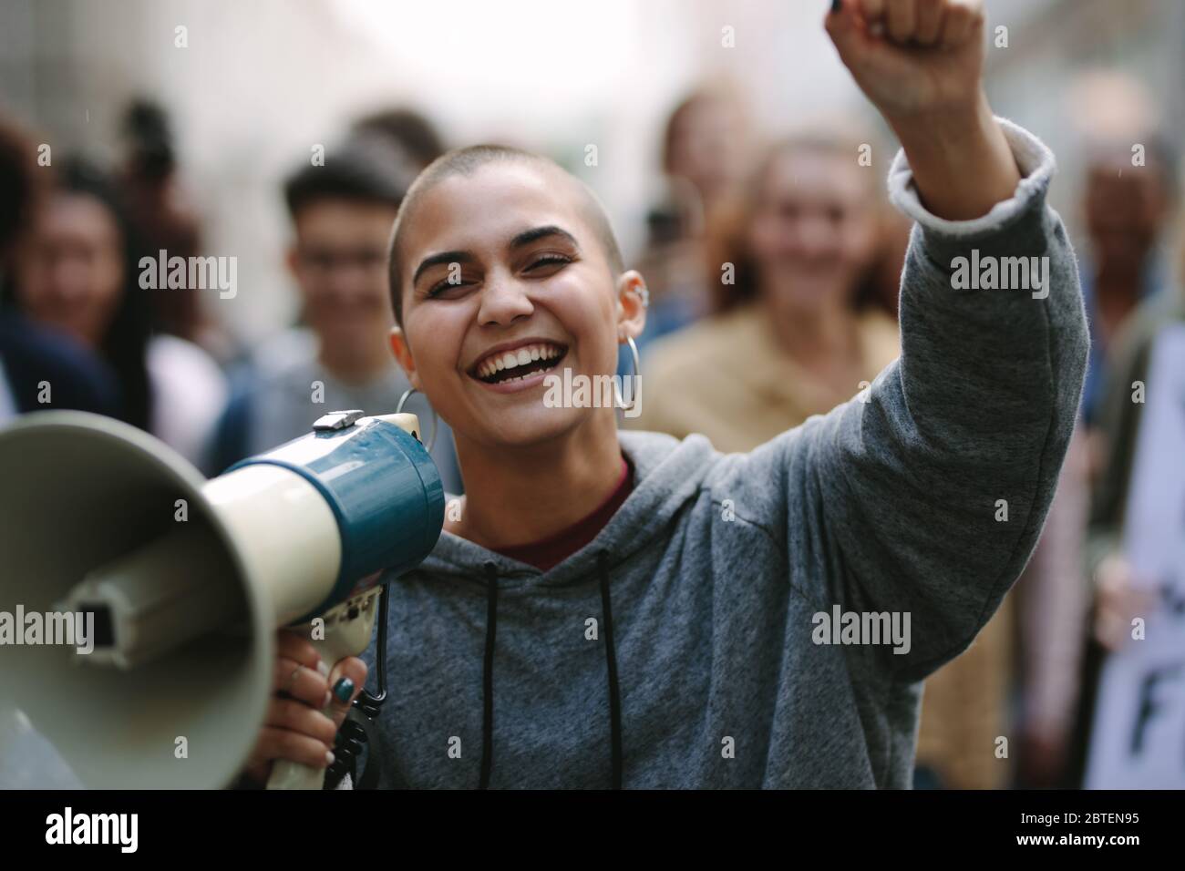Lächelnde Frau protestiert mit einem Stierkampfhorn auf der Straße mit Menschen im Hintergrund. Weiblich mit einem Megaphon, da sie an einer Straßendemonstratio teilnimmt Stockfoto
