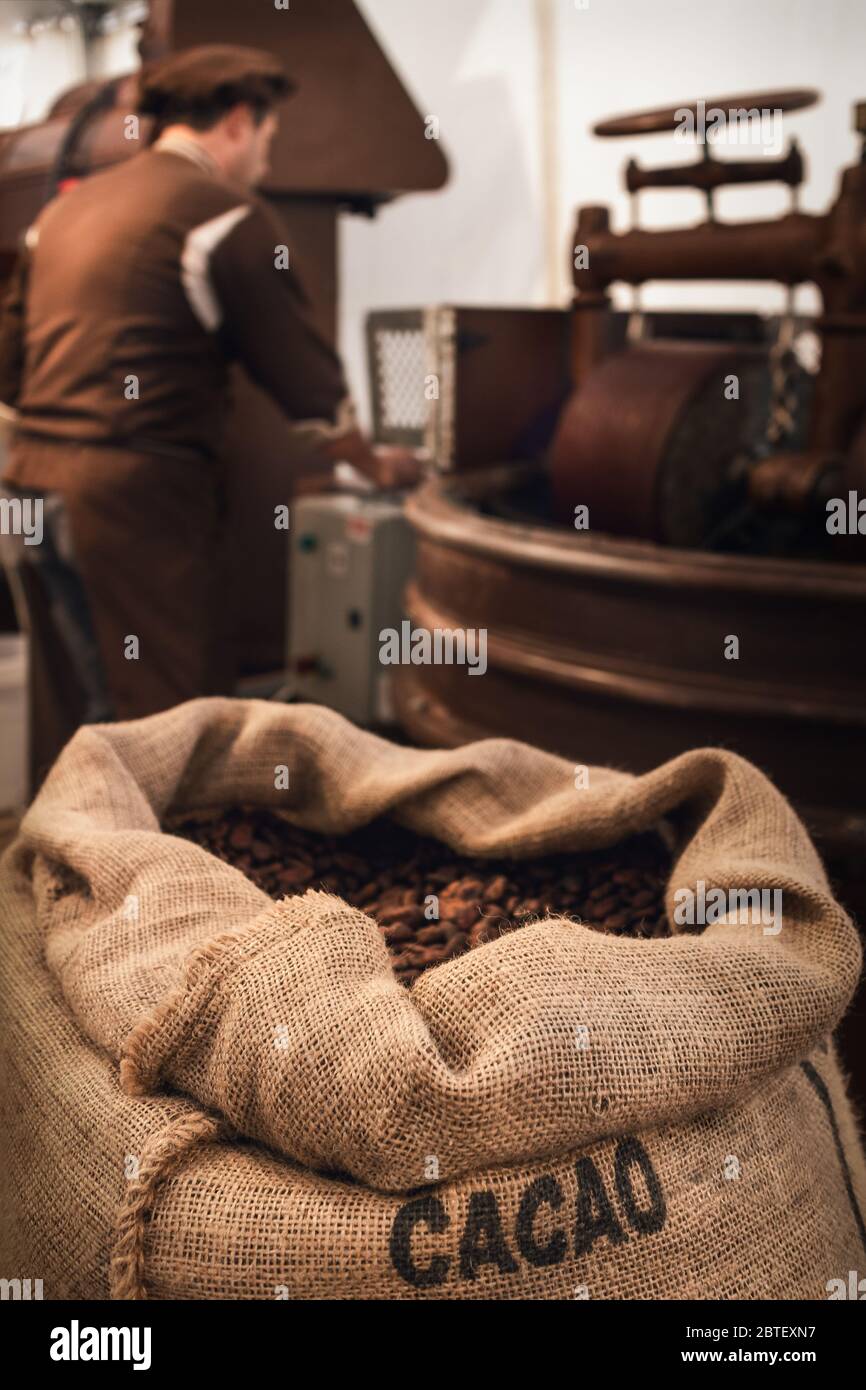 Jute Beutel voller Kakaobohnen in einer Schokoladenfabrik, mit einem männlichen Chocolatier arbeiten an Conchieren und Melanger Ausrüstung auf dem Hintergrund Stockfoto