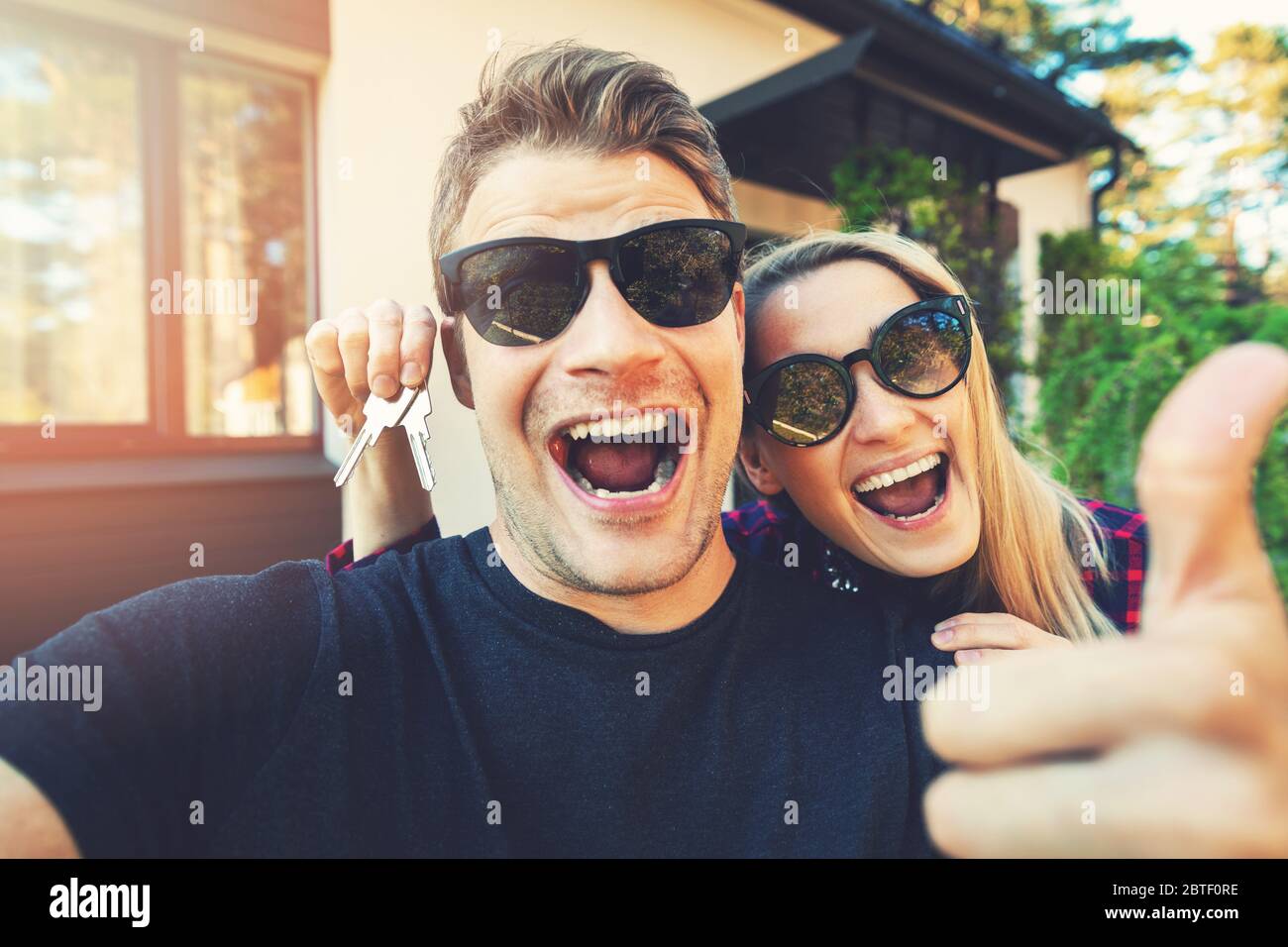 Junge glückliche Paar mit Schlüsseln in der Hand stehen außerhalb ihres neuen Hauses und machen ein Selfie Stockfoto