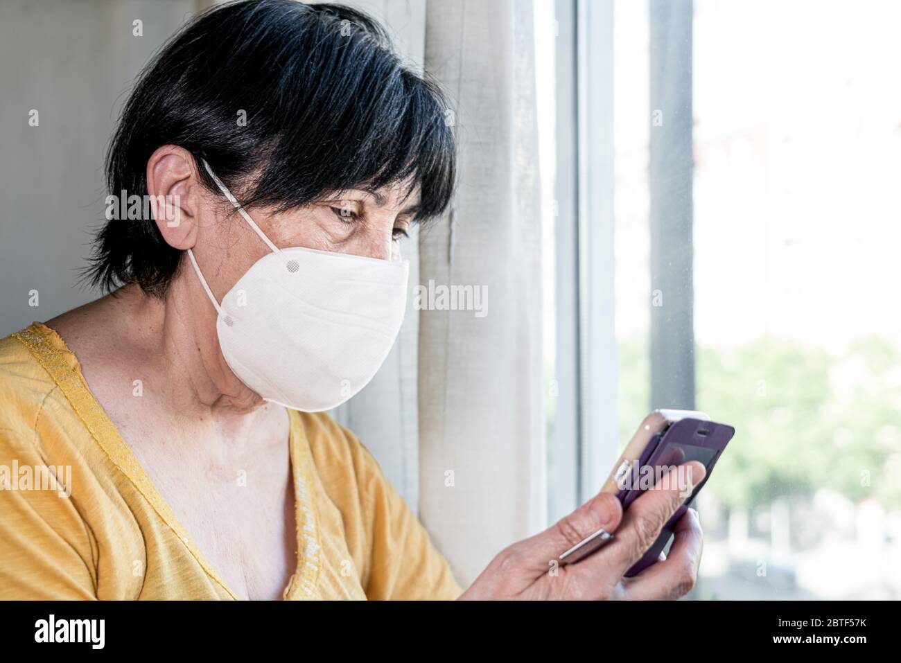 Alte Frau glücklich das Smartphone mit Gesichtsmaske im Fenster suchen. Barcelona, Spanien. Stockfoto Alte Frau glücklich das Smartphone mit Gesichtsmaske im Fenster suchen. Barcelona, Spanien. Stockfoto