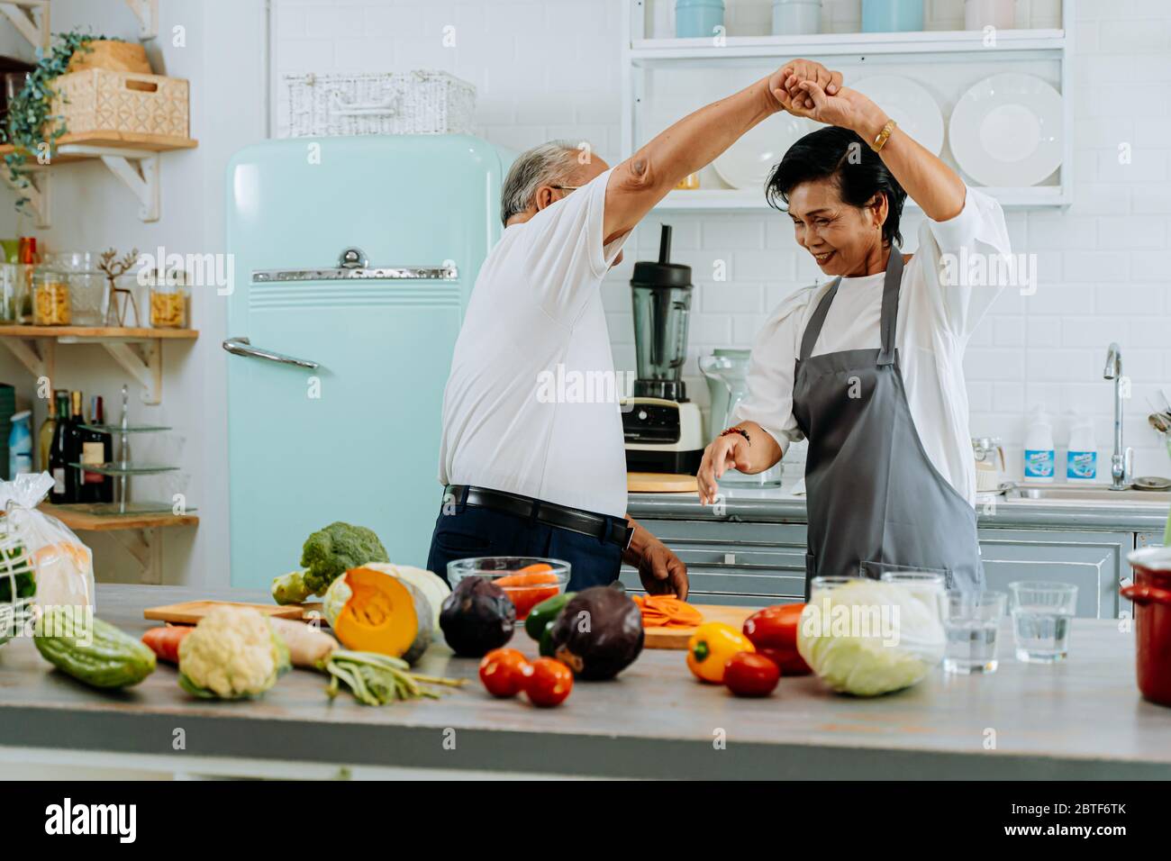 Asiatische ältere Paar tanzen und lächeln in der Küche. 70er Jahre älterer Mann und Frau kochen zusammen zu Hause. Stockfoto