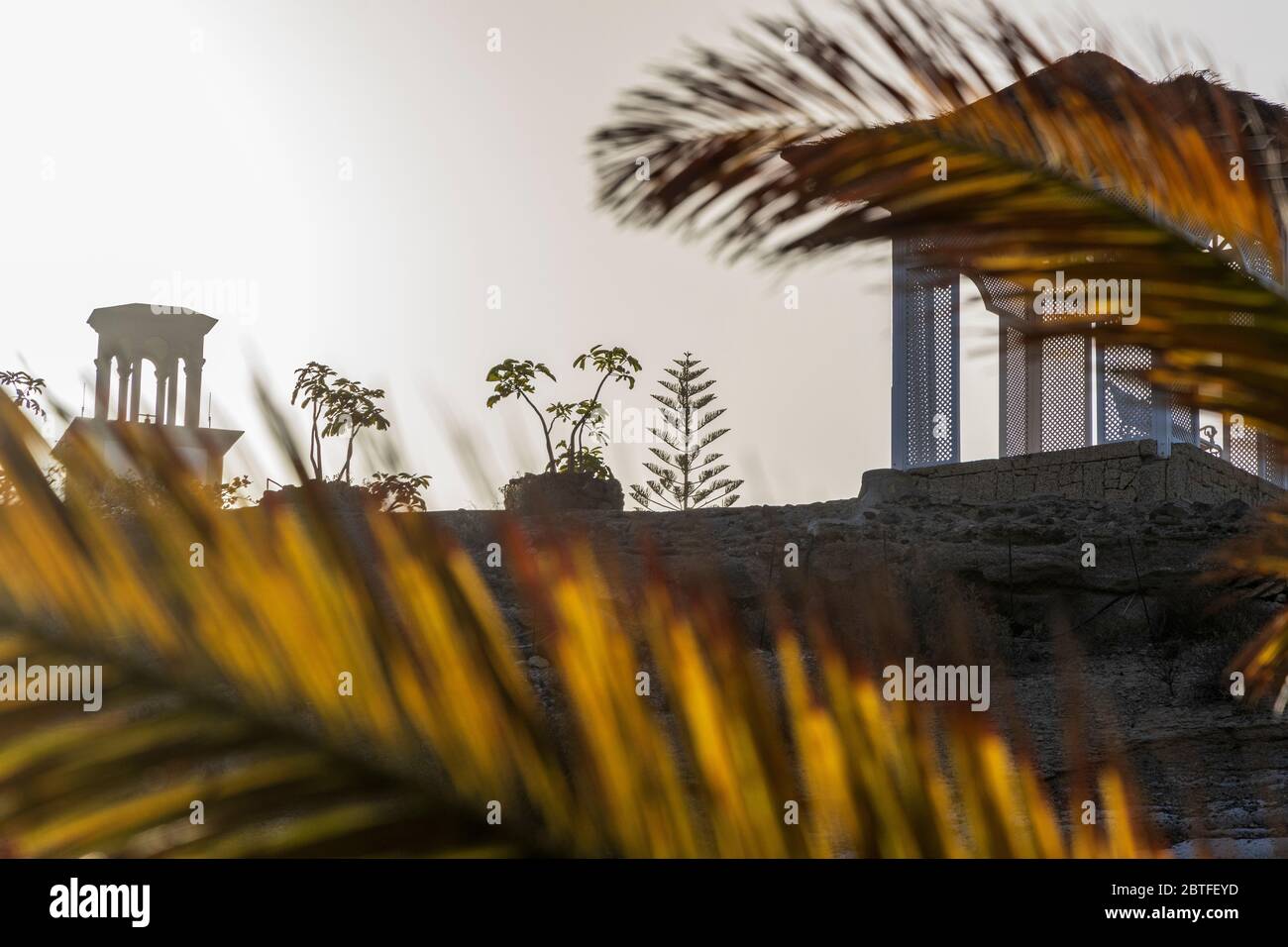 Blick durch vergilbte Palmenblätter auf den hinterleuchteten Pavillon auf einem Kamm über dem Strand Playa del Duque am ersten Tag der zweiten Phase der Deeskalation, Stockfoto