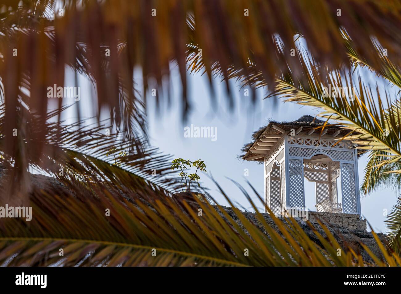 Blick durch vergilbte Palmenblätter auf den hinterleuchteten Pavillon auf einem Kamm über dem Strand Playa del Duque am ersten Tag der zweiten Phase der Deeskalation, Stockfoto