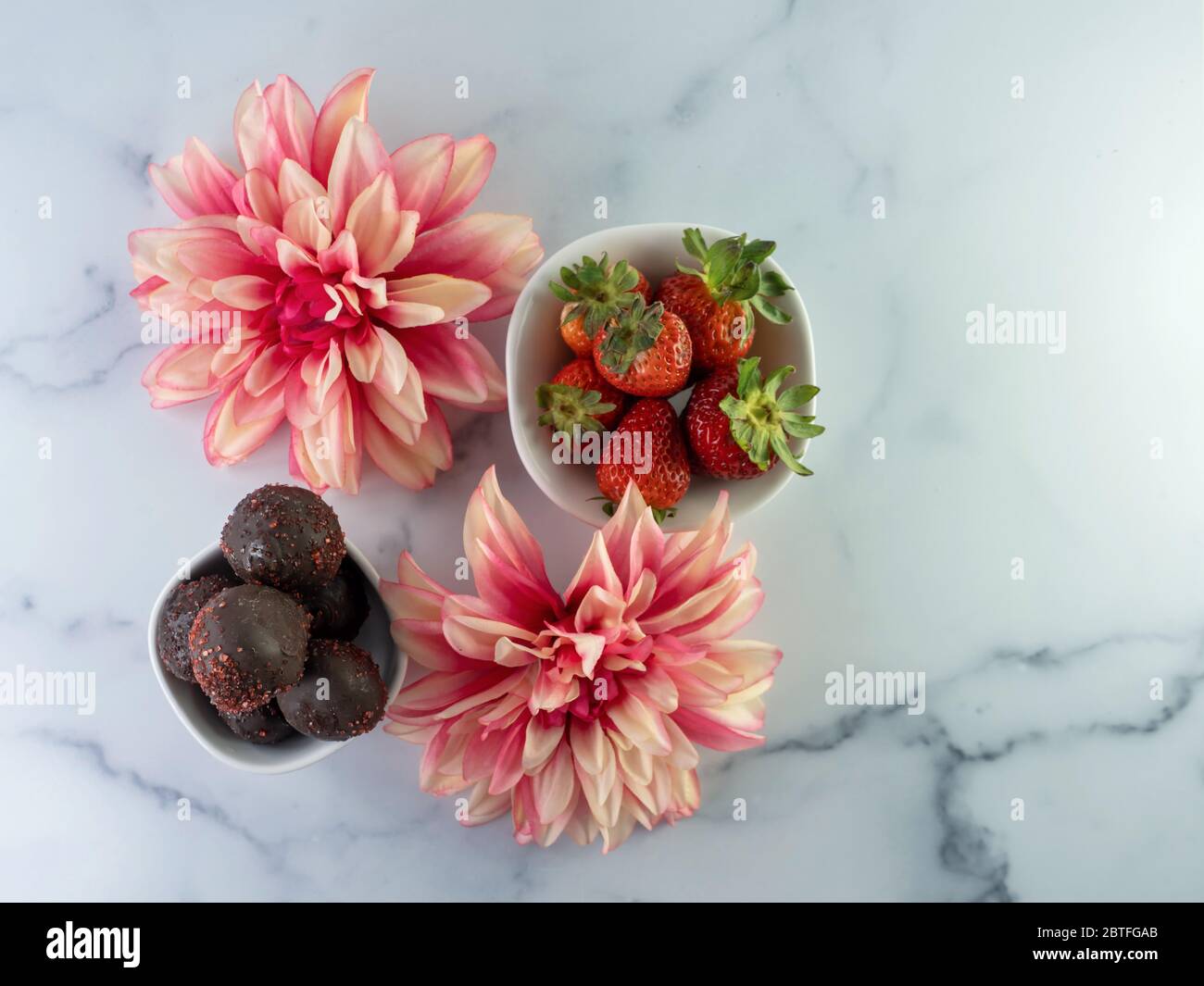 Flach legen Fotografie einer Schüssel voller Erdbeeren und eine Schüssel voll von Schokolade bedeckt Erdbeerkuchen Kugeln mit zwei rosa Dahlia Kunstblume Stockfoto