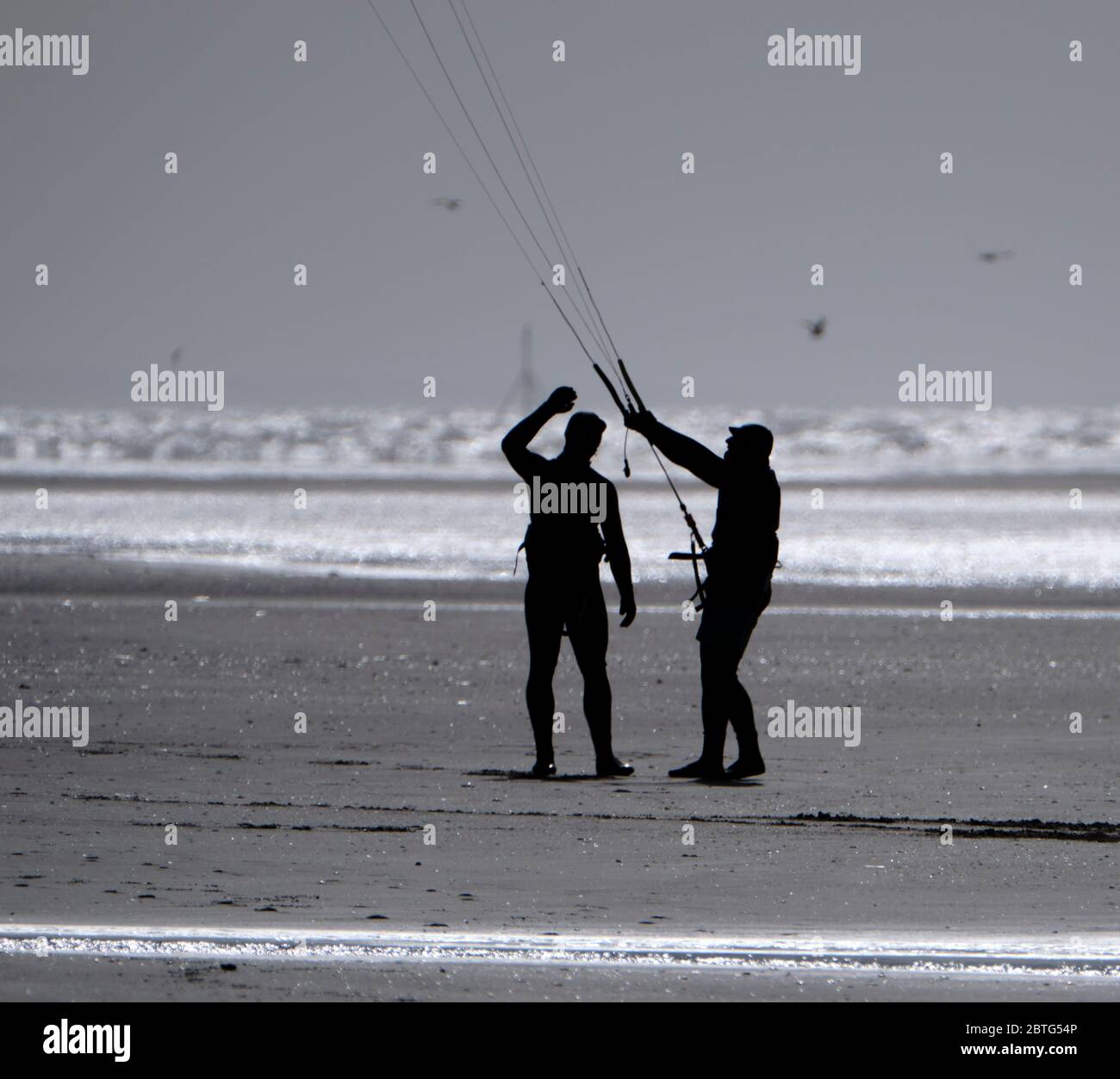 Silhouette von zwei Personen mit einem Drachen am Strand, vor einem hellen Himmel, der auf nassem Sand reflektiert. Stockfoto