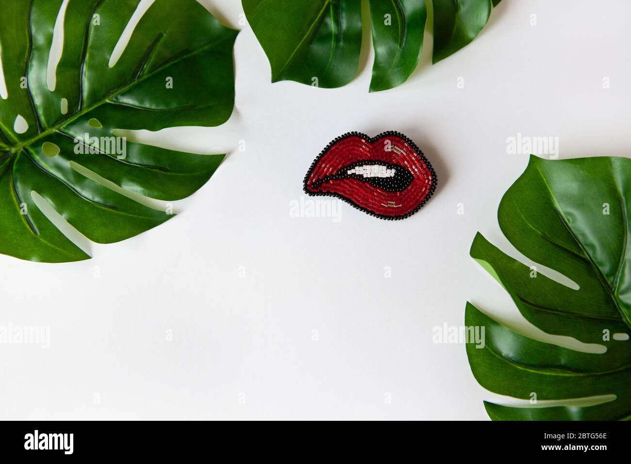 Modische Brosche in Form von Lippen aus japanischen Perlen, grüne Blätter von Monstera auf weißem Hintergrund. Nahaufnahme, flaches Layout, Kopierbereich. Konzepttrends, Stockfoto