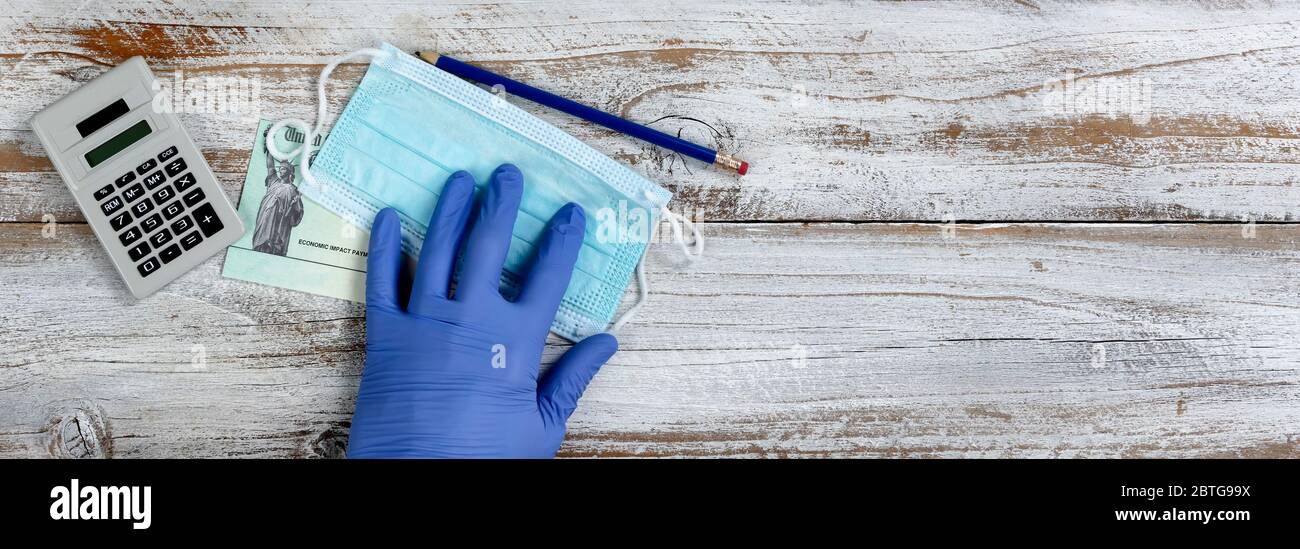 Handgriff mit Handschuhen für persönliche Schutzmaske mit wirtschaftlichem Impact Stimulus Check auf weißem Holztischunterlage Stockfoto