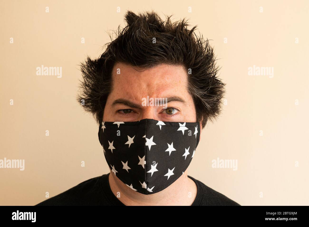 Ein Mann in seinen späten dreißiger Jahren trägt wiederverwendbare Gesichtsmaske und schaut direkt auf den Betrachter während der Coronavirus-Pandemie Covid 19 mit wildem, absperrbarem Haar Stockfoto