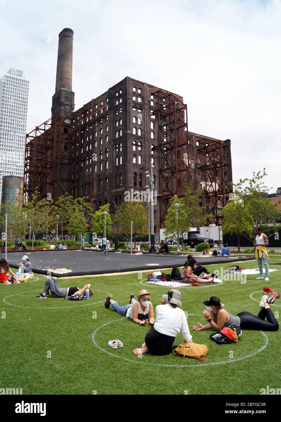 Menschen, die Gesichtsmasken tragen, sich auf einem Gras im Domino Park entspannen, im Hintergrund das ehemalige Zuckerraffiniergebäude in Brooklyn, NY, USA Stockfoto