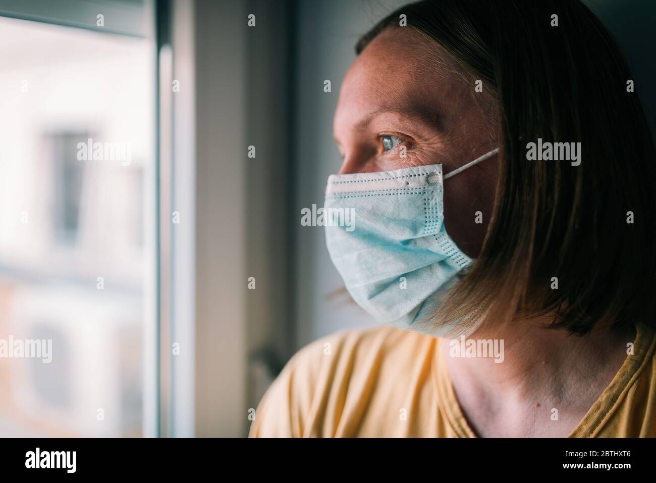 Frau in Selbstisolation beim Virusausbruch, die durch Fenster schaut. Besorgte weibliche Person mit chirurgischer Schutzmaske und selektivem Fokus Stockfoto