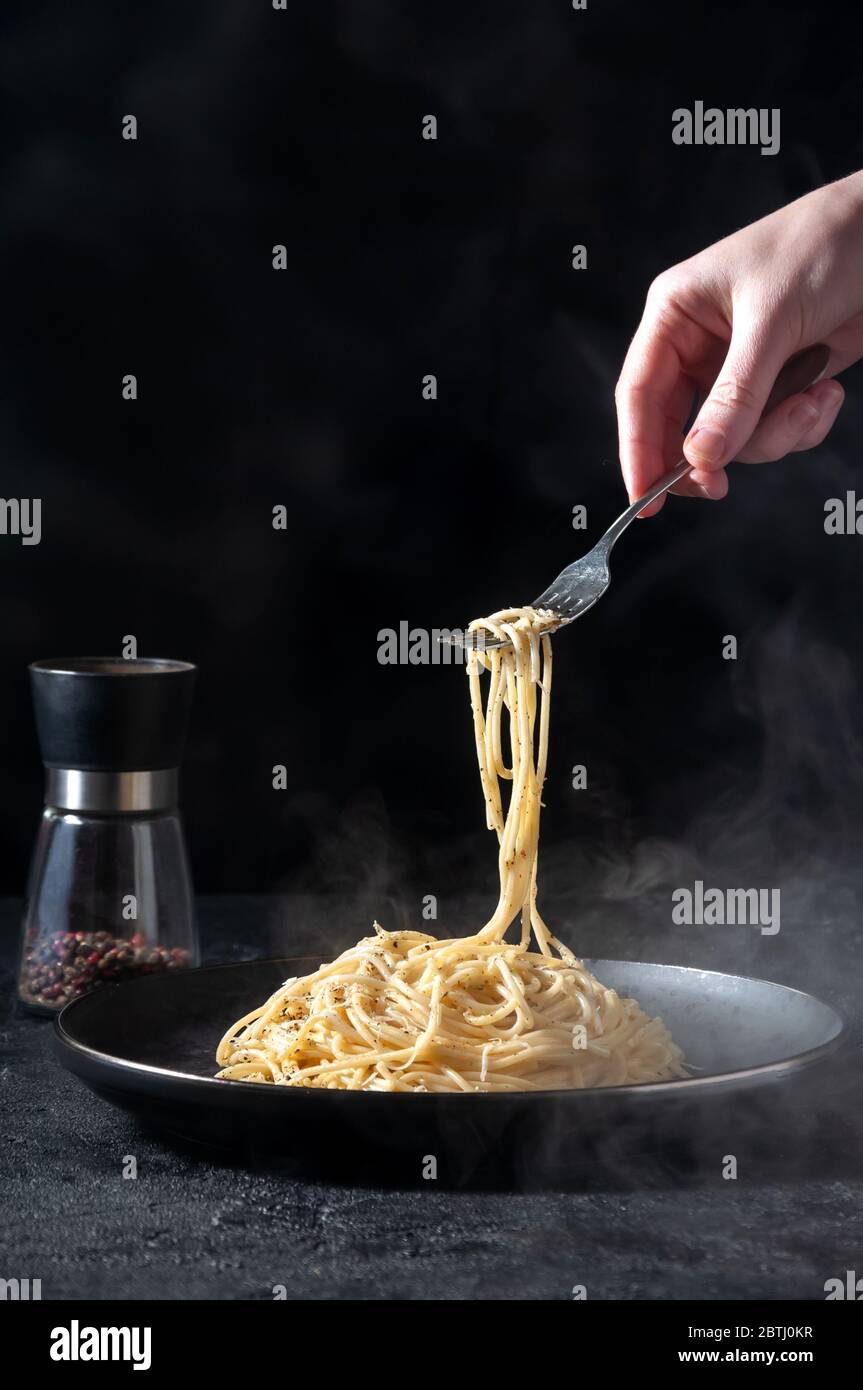 Cacio e Pepe - heiße italienische Pasta mit Käse und Pfeffer auf schwarzem Teller, Frau, die Gabel mit Spaghetti auf dunklem Hintergrund hält. Stockfoto