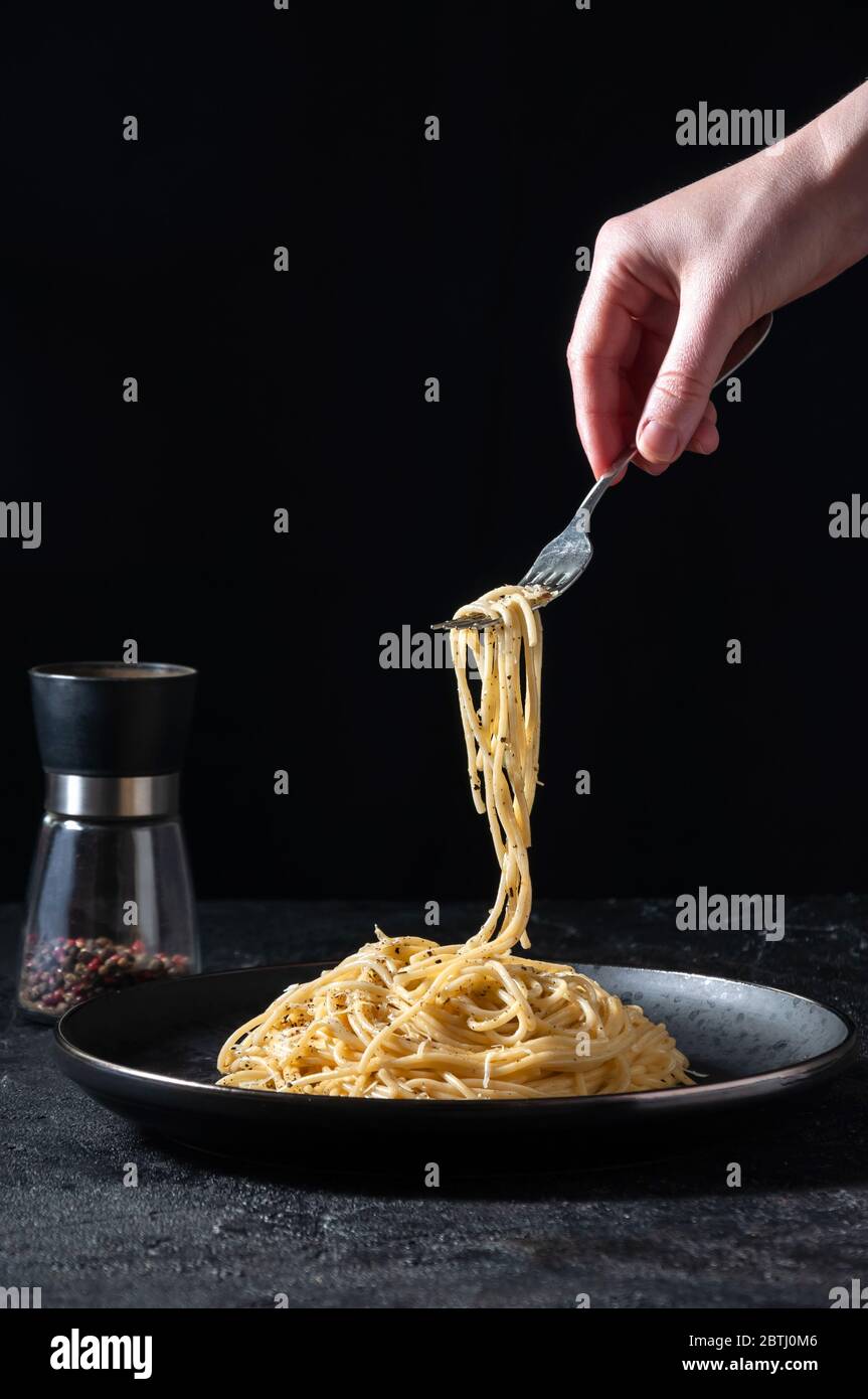 Cacio e Pepe - heiße italienische Pasta mit Käse und Pfeffer auf schwarzem Teller, Frau, die Gabel mit Spaghetti auf dunklem Hintergrund hält. Stockfoto