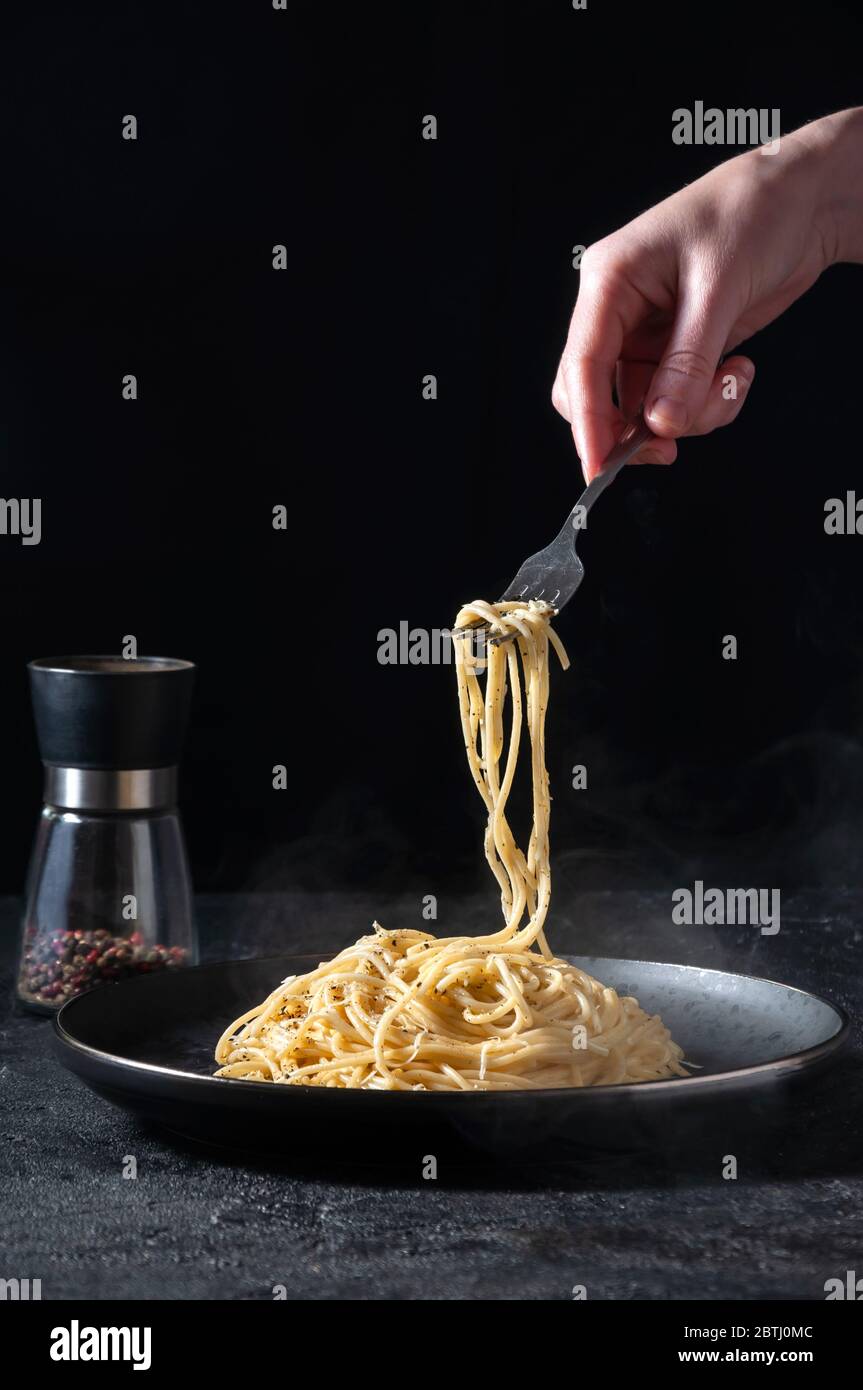 Cacio e Pepe - heiße italienische Pasta mit Käse und Pfeffer auf schwarzem Teller, Frau, die Gabel mit Spaghetti auf dunklem Hintergrund hält. Stockfoto