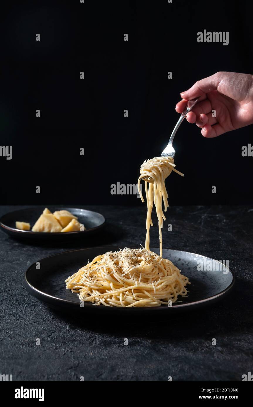 Cacio e Pepe - heiße italienische Pasta mit Käse und Pfeffer auf schwarzem Teller, Frau, die Gabel mit Spaghetti auf dunklem Hintergrund hält. Stockfoto