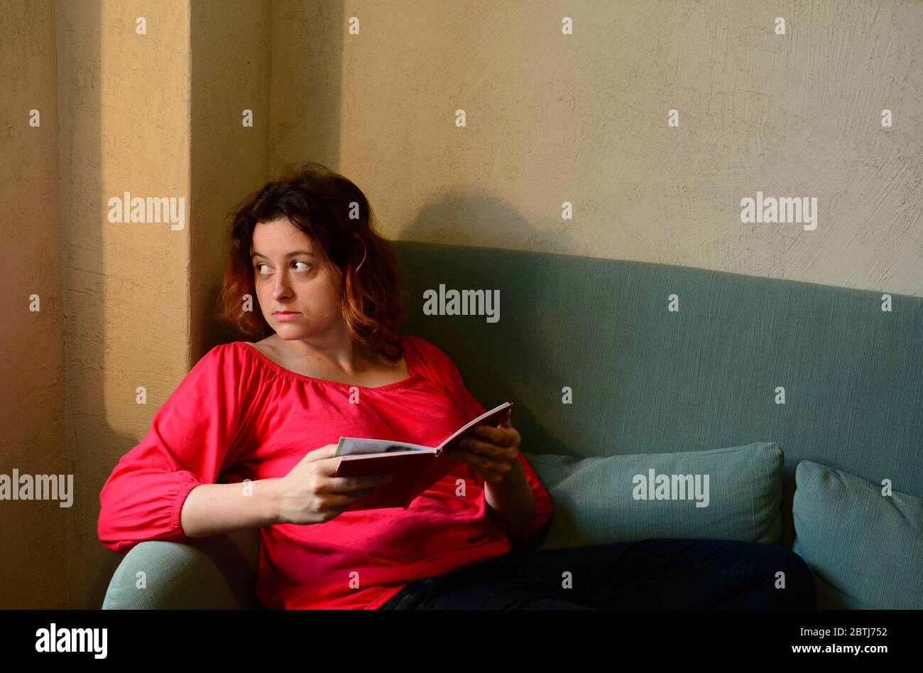 Junge Frau, die mit einem Buch auf einem Sofa im Wohnzimmer sitzt. Zu Hause bleiben während der Isolationszeit und Quarantänekonzept Stockfoto