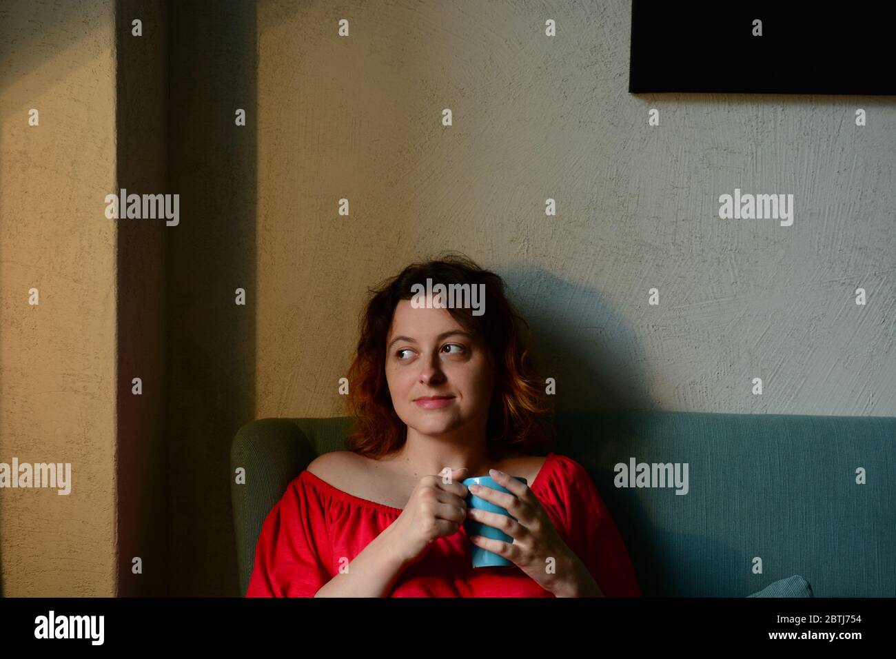 Junge Frau sitzt auf einem Sofa im Wohnzimmer und genießt Kaffee. Zu Hause bleiben während der Isolationszeit und Quarantänekonzept Stockfoto