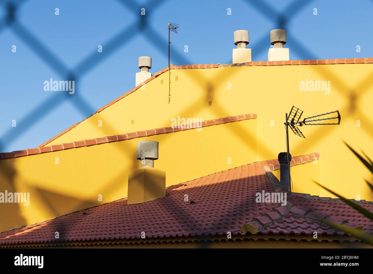 Sonne auf gelben Wänden und Terrakotta-Fliesen der örtlichen Schule durch einen Zaun gegen blauen Himmel während der zweiten Phase der Deeskalation des Staates eme Stockfoto