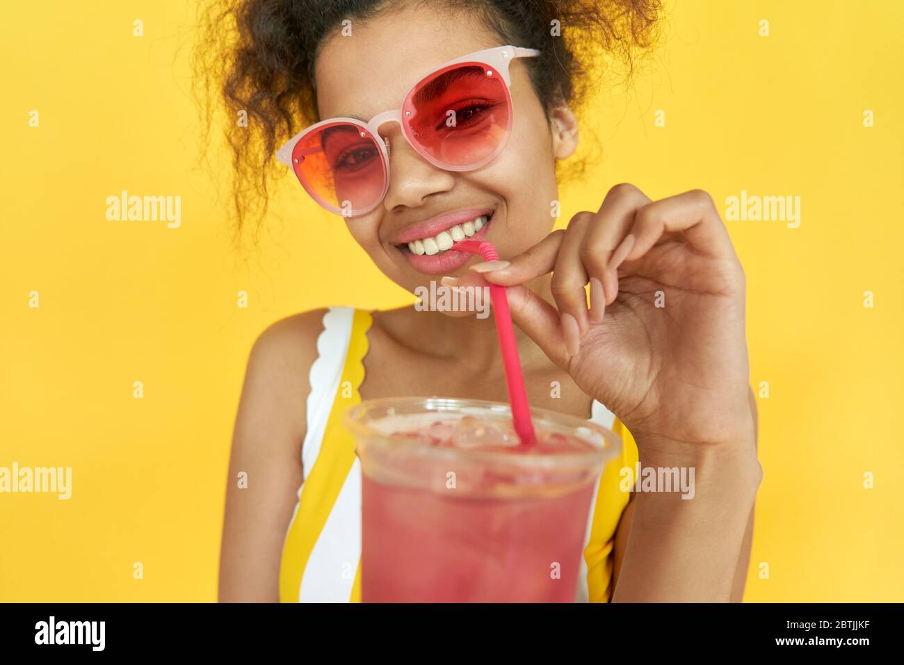 Lächelnd afrikanischen Teenager Mädchen trinken Cocktail tragen Sonnenbrille isoliert auf gelb. Stockfoto