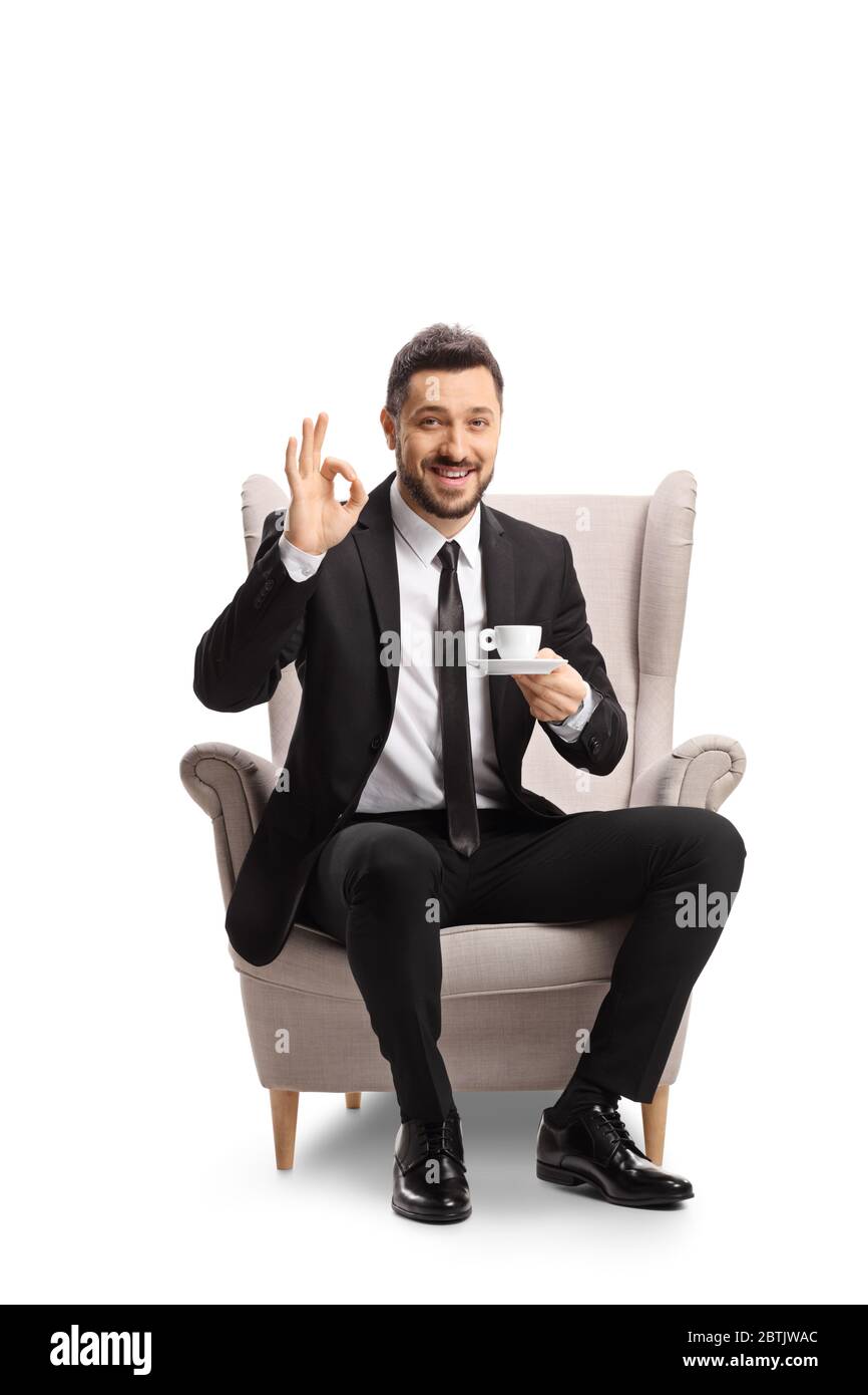 Junger Mann in Anzug und Krawatte sitzt in einem Sessel mit einer Tasse Espresso Kaffee und gesturing ok Zeichen isoliert auf weißem Hintergrund Stockfoto