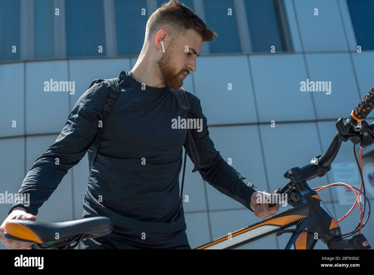 Bärtiger Radfahrer junger Mann in kabellosen Kopfhörern, vor dem Hintergrund der Wolkenkratzer eines Bürogebäudes. Im Freien. Frühlingssport. Der Typ ist es Stockfoto