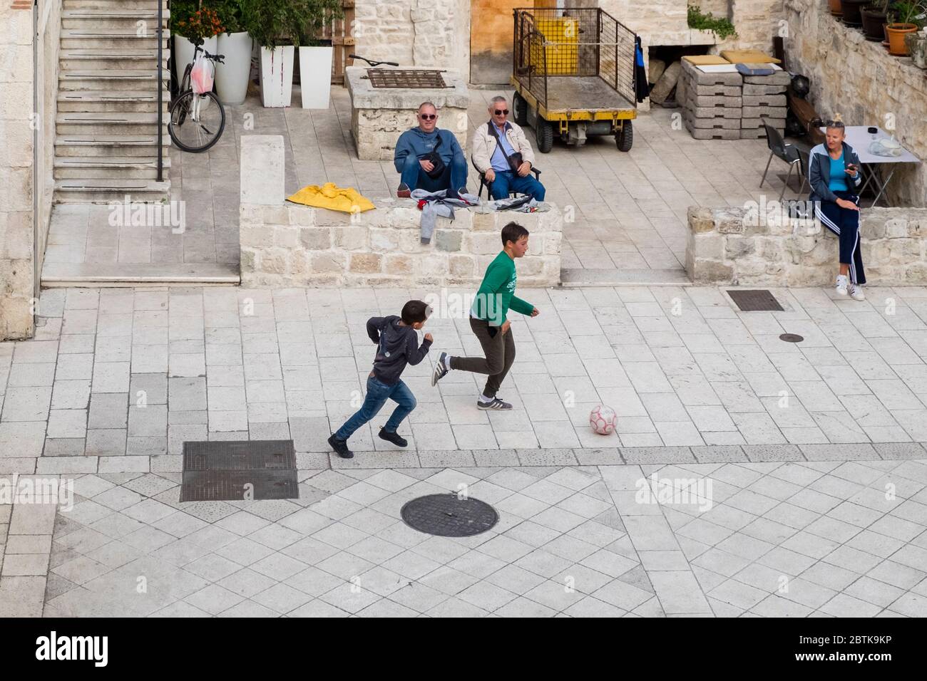 Zwei kroatische Jungen spielen Fußball, während drei kroatische Erwachsene beobachten, in einem Wohnhof, Altstadt, Split, Kroatien Stockfoto
