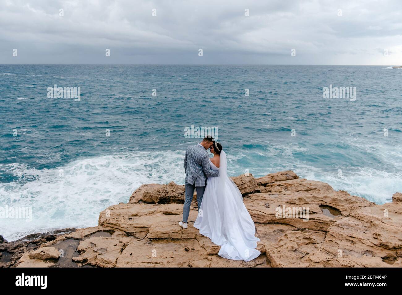 Das Brautpaar umarmt sanft die Felsen am Meer und genießt die Natur Zyperns. Rückansicht. Flitterwochen des Brautpaids Stockfoto