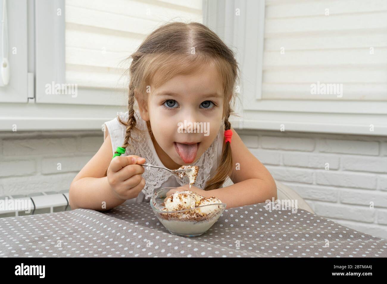 Ein 4-jähriges Mädchen in einer weißen Bluse isst gerne Eis mit Schokolade aus den Töpfen am Tisch in der Küche. Das Kind öffnet mit Stockfoto