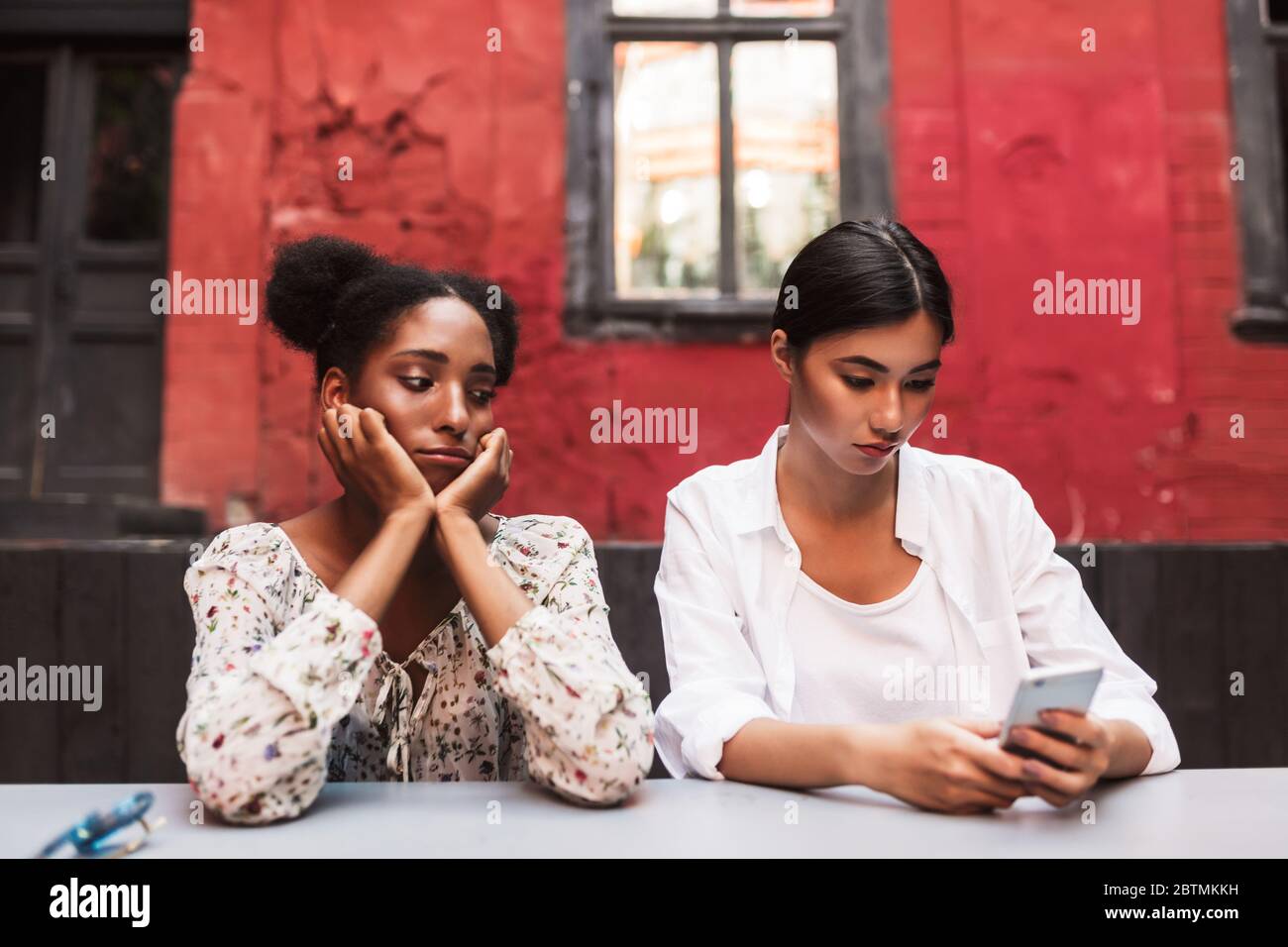 Aufgeregt african Mädchen traurig beiseite schauen, während asiatische Mädchen in der Nähe nachdenklich mit Handy im Innenhof des Cafés Stockfoto