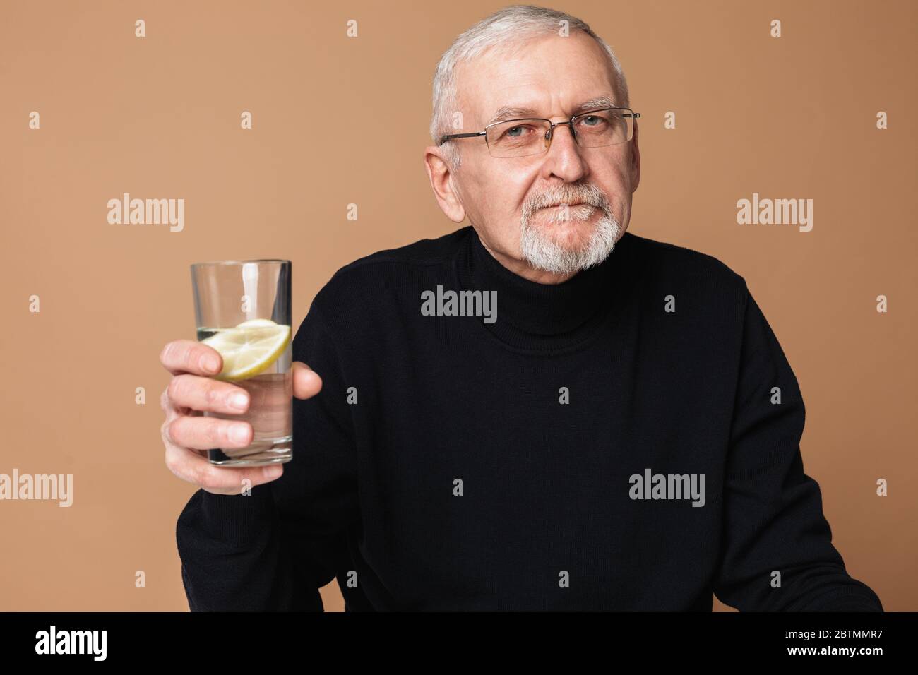 Alter schöner Mann mit grauen Haaren und Bart in Brillen und Pullover nachdenklich in der Kamera halten Glas von reinem Wasser mit einer Scheibe Zitrone Stockfoto