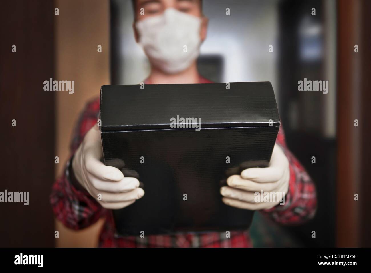 Nach Hause liefern, Online-Bestellung. Mann in einer medizinischen Maske und Gummihandschuhe mit schwarzer Box, ein Paket in den Händen. Lieferung während der Quarantäne der Stockfoto