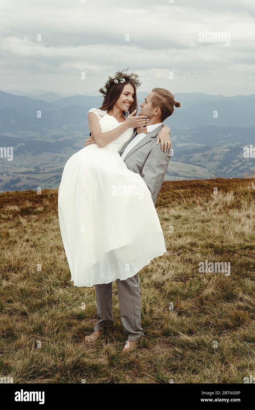 Stilvolle Bräutigam Holding glücklich Braut in Sonne Licht und lächelnd, romantischen Moment, Boho Hochzeit Paar, Luxus-Zeremonie in den Bergen mit erstaunlichen V Stockfoto
