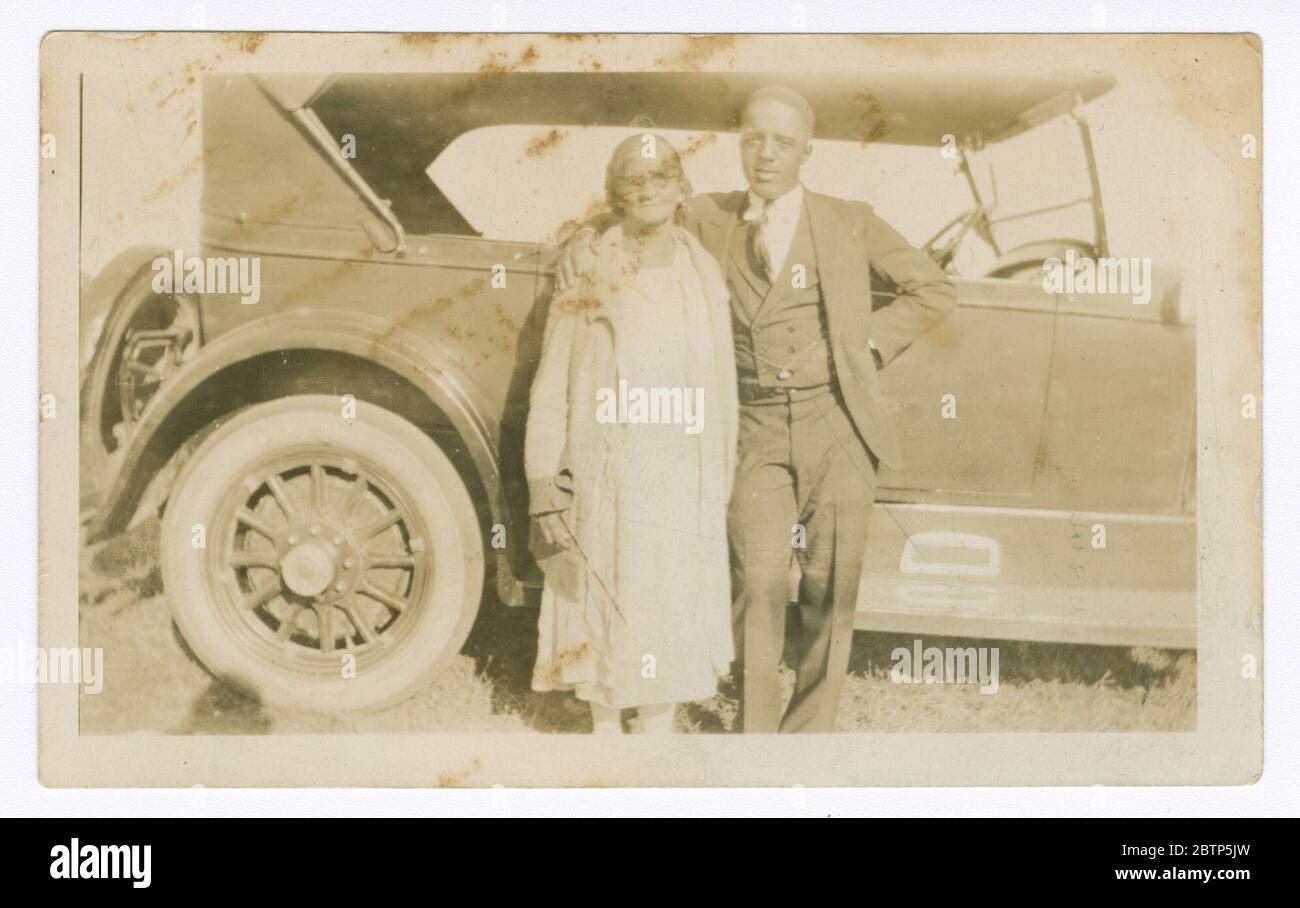 Foto eines Mannes und einer Frau vor dem Auto. Ein Schwarz-Weiß-Foto eines Mannes und einer Frau, die vor einem Auto stehen. Der Mann trägt einen dreiteiligen Anzug und wickelt seine Arme um die Schultern der Frau, die ein Kleid und eine Jacke trägt. Auf der Rückseite ist [34] eingestanzt. Stockfoto