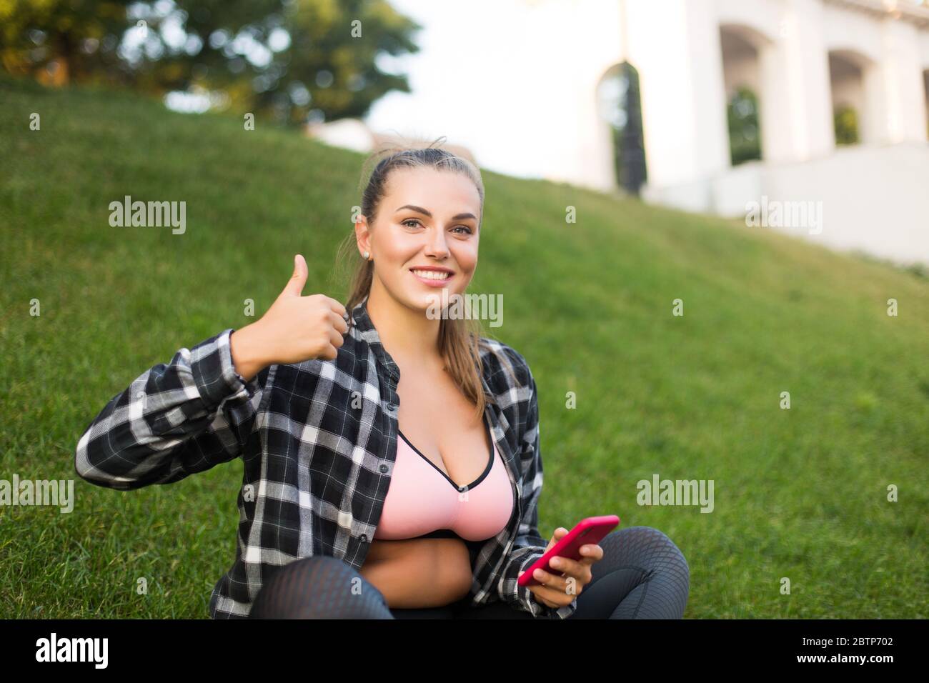 Junge hübsche plus size Frau in lässigem Hemd und sportlichem Oberteil, das Handy in der Hand hält, glücklich in der Kamera schauend, die große Daumen-nach-oben-Geste zeigt Stockfoto