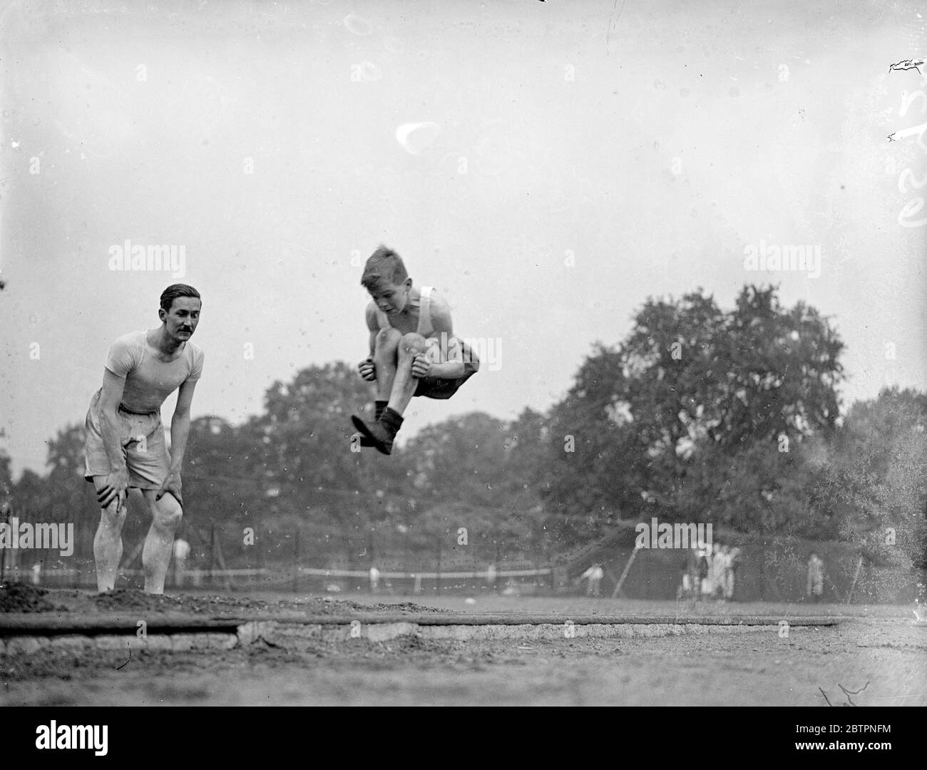Eine Lektion von Papa Frederik Robert Miller, ehemaliger walisischer Rugby-Nationalspieler, der heute Sportmeister an der St. Johns School in Brixton ist und seinem Sohn zeigt, wie man im Battersea Park in London Weitsprung macht. 19. Juni 1937 Stockfoto