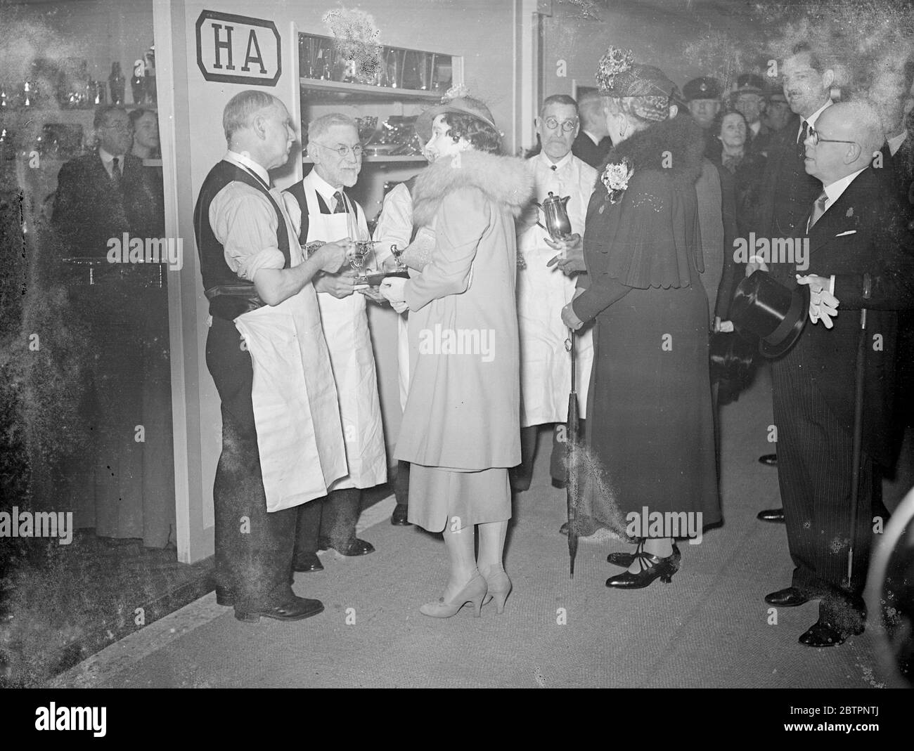 Königin und Königin Mary, sprechen Sie mit dem alten Crossman im Olympia BIF. Die Königin und Königin Mary besuchten wieder die Olympia-Sektion der British Industries Fair und unterhielten sich während ihrer Tour mit vier alten Sheffield-Handwerkern, die dazu beitrugen, den Silbersalver dem König von der Sheffield Chamber of Commerce und der Cutler's Company zu überbringen. Zwischen ihnen haben die vier Männer 241 Jahre lang bei den Atkin Brothers, den Sheffield Silberschmieden, gearbeitet. Es sind Harry Dennis, Metal, Smith (63 Jahre Dienst), Jack Stokes, Silberschmiede (62 Jahre Dienst), Ezra Lee, Silberschmiede (60 Jahre) und Sidney Jon Stockfoto