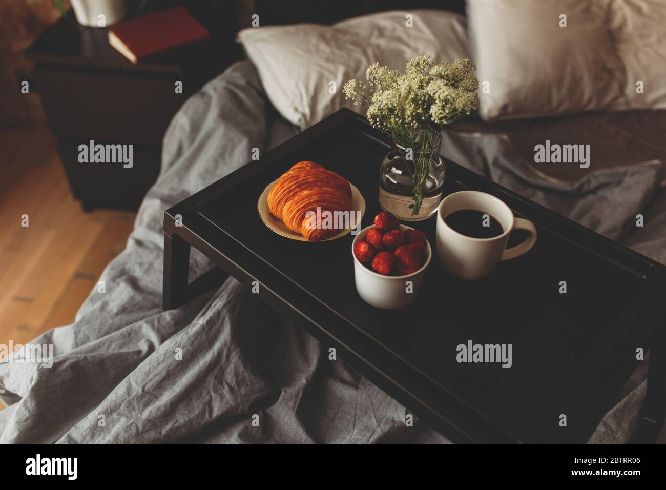 Frühstück auf einem Tablett im Bett zu Hause weiße dunkle Bettwäsche. Croissant Tee Erdbeeren. Stockfoto