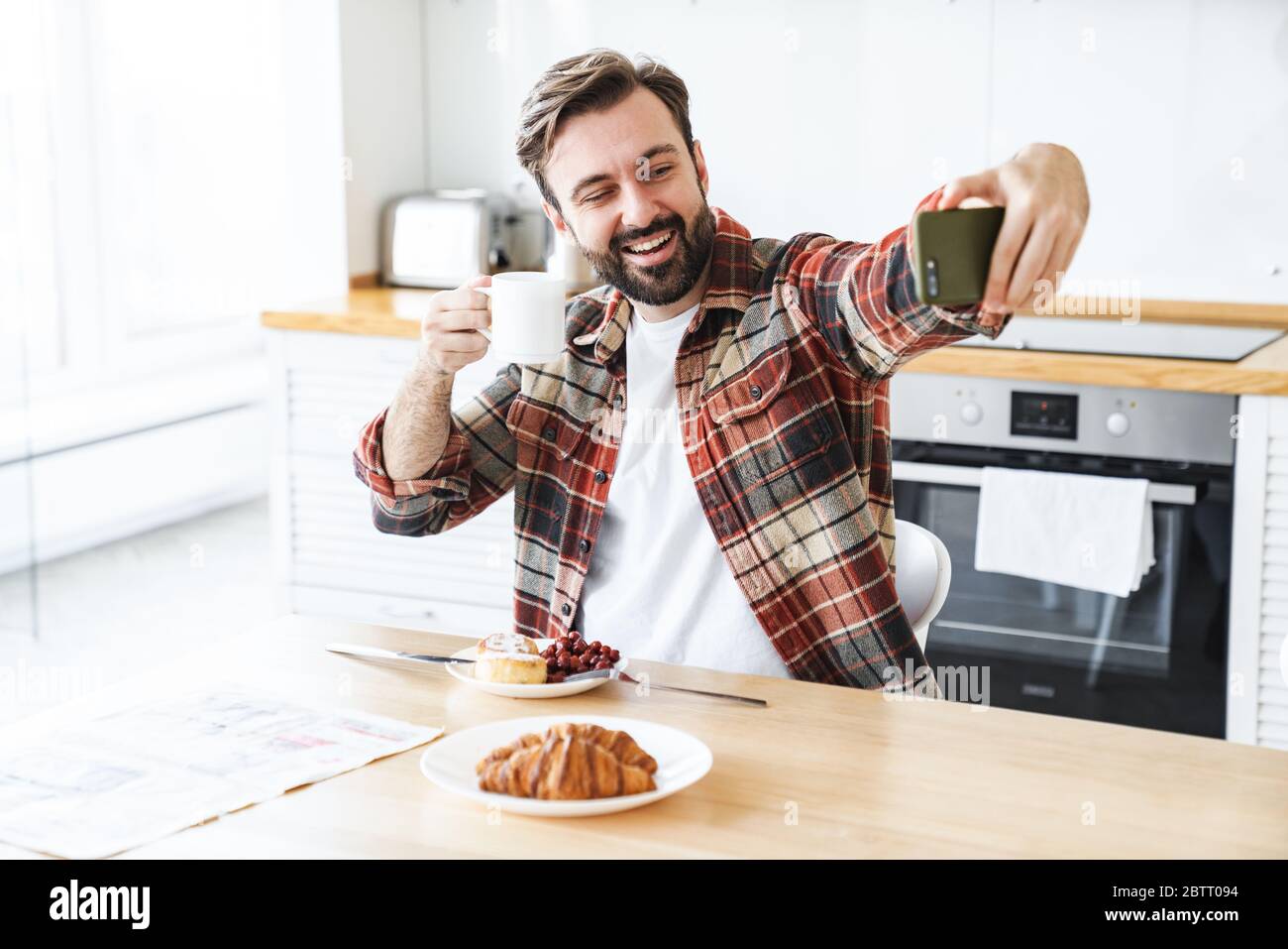 Porträt eines lächelnden bärtigen Mannes, der Selfie auf dem Handy nimmt und Kaffee trinkt, während er zu Hause frühstücken kann Stockfoto