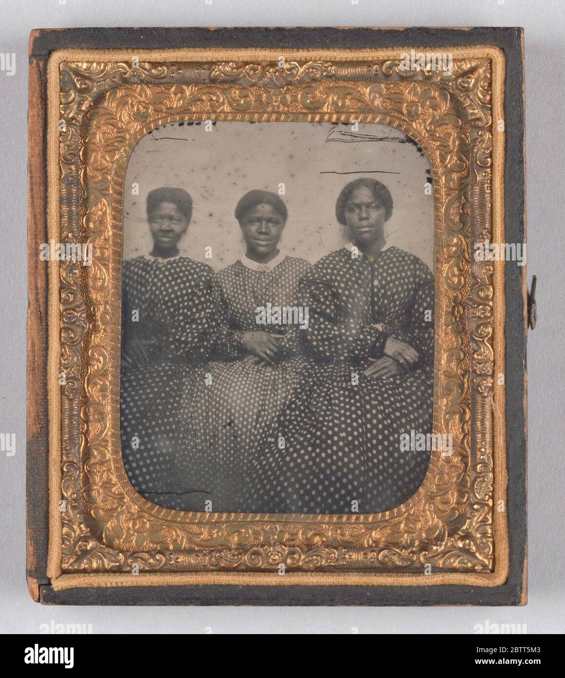 Ambrotype von drei Frauen in gepunkteten calico Kleidern. Ein Schwarz-Weiß-Foto von drei Frauen, die in einer Reihe sitzen, die Arme an den Ellbogen miteinander verbunden und die Hände in ihren Schoß von rechts über links gelegt haben. Alle drei Frauen tragen Kleider aus dunklem Stoff mit hellen Tupfen. Stockfoto