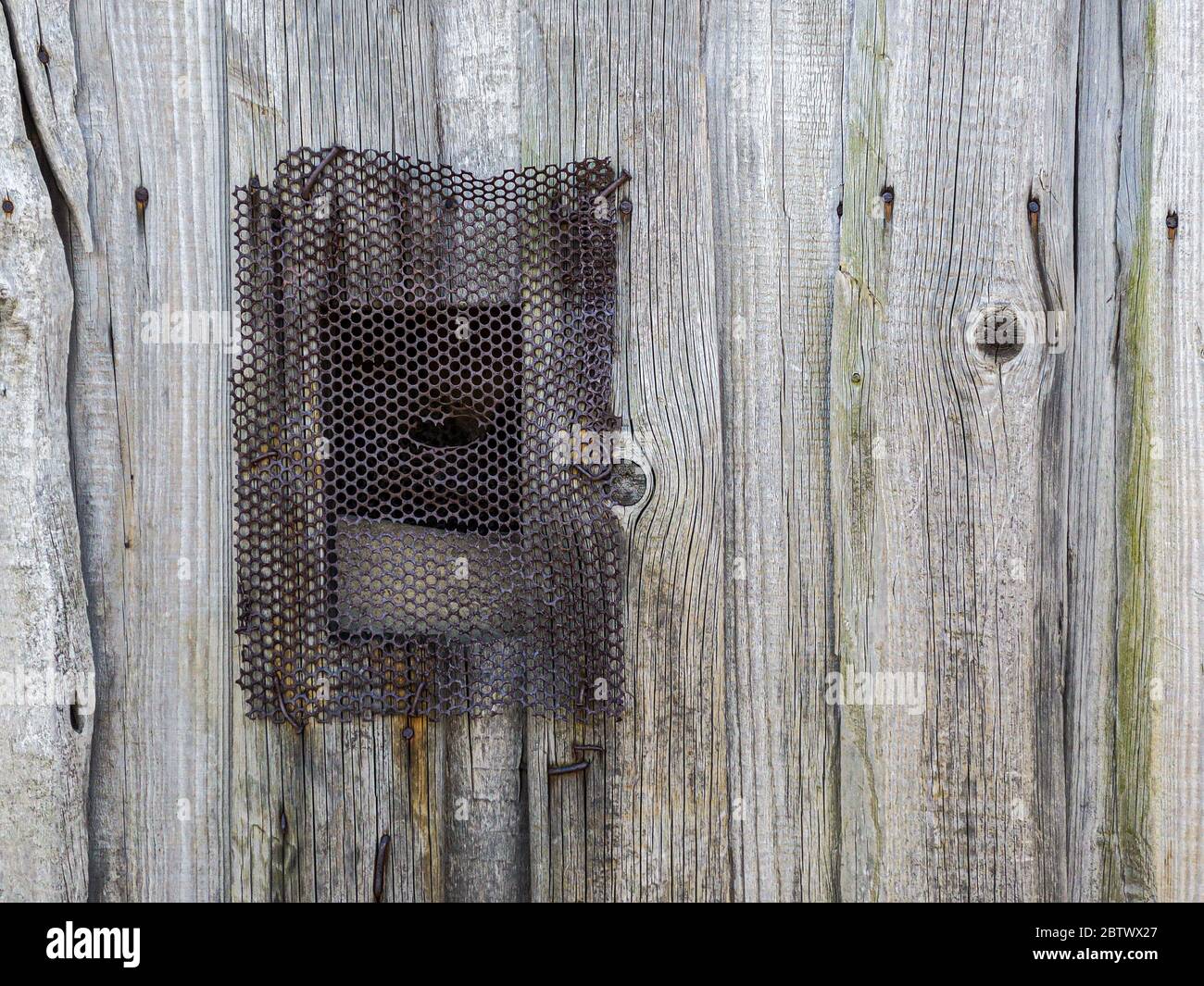 Kleines Fenster mit perforiertem rostigen Metallrost auf grau verwitterter Holzwand Stockfoto