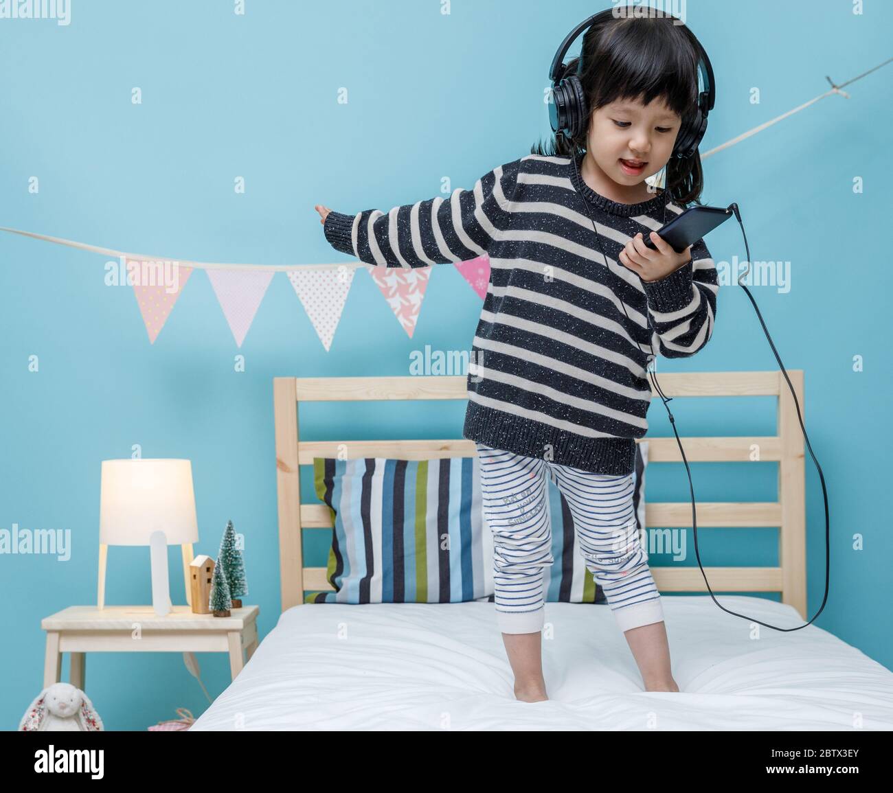 Nettes kleines Mädchen singen ein Lied mit Smartphone in ihrem Schlafzimmer, glücklich asiatische Kind kleines Mädchen, das die Musik mit Kopfhörer auf dem Bett, Technologie c Stockfoto