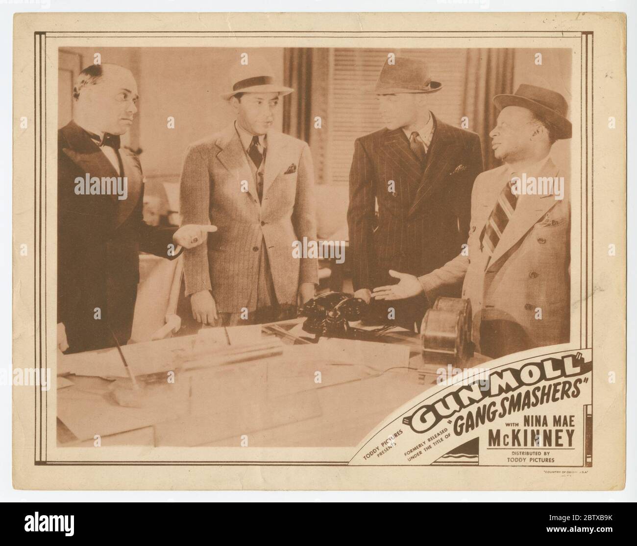 Lobby-Karte für Gun Moll aka Gun Smashers. Eine Lobby-Karte für den Film 'Gun Moll'. Die Karte besteht aus einem Schwarz-Weiß-Bild von vier Männern, die um einen Schreibtisch in einem Büro stehen und sprechen. Drei der Männer tragen Anzüge und Hüte und ein Mann trägt einen Tux. Stockfoto